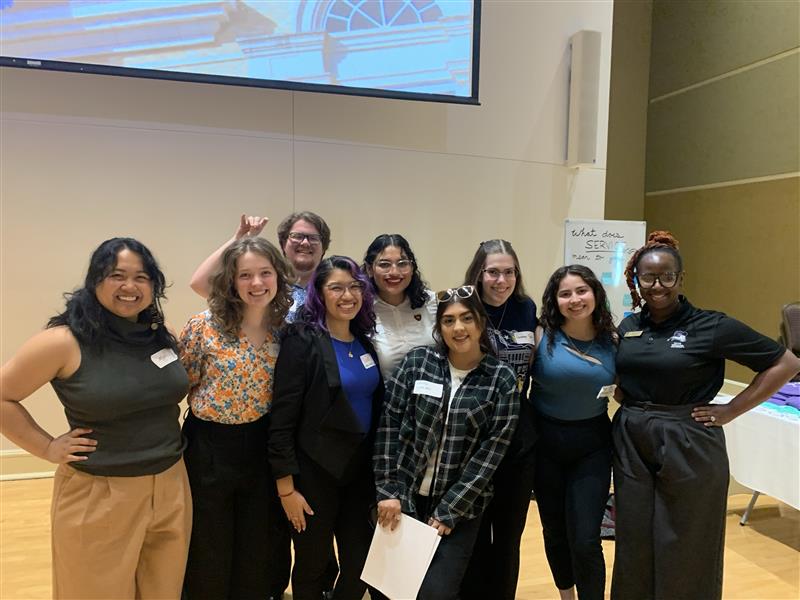 Group of eight women standing together indoors, smiling, some holding papers, in a conference room setting.