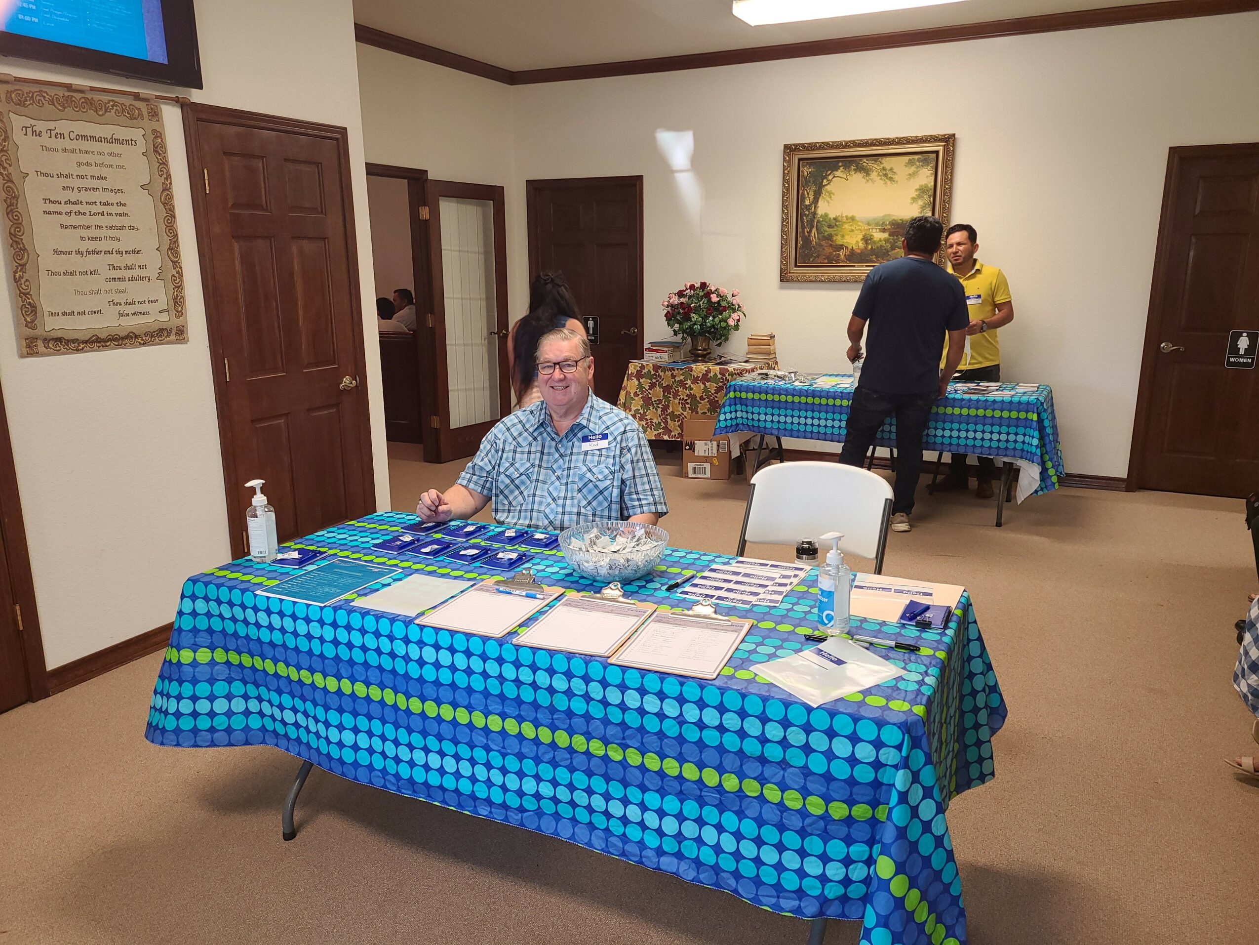 Man sitting at a table with a blue checkered tablecloth, papers, and a water bottle in a room with other tables and people.
