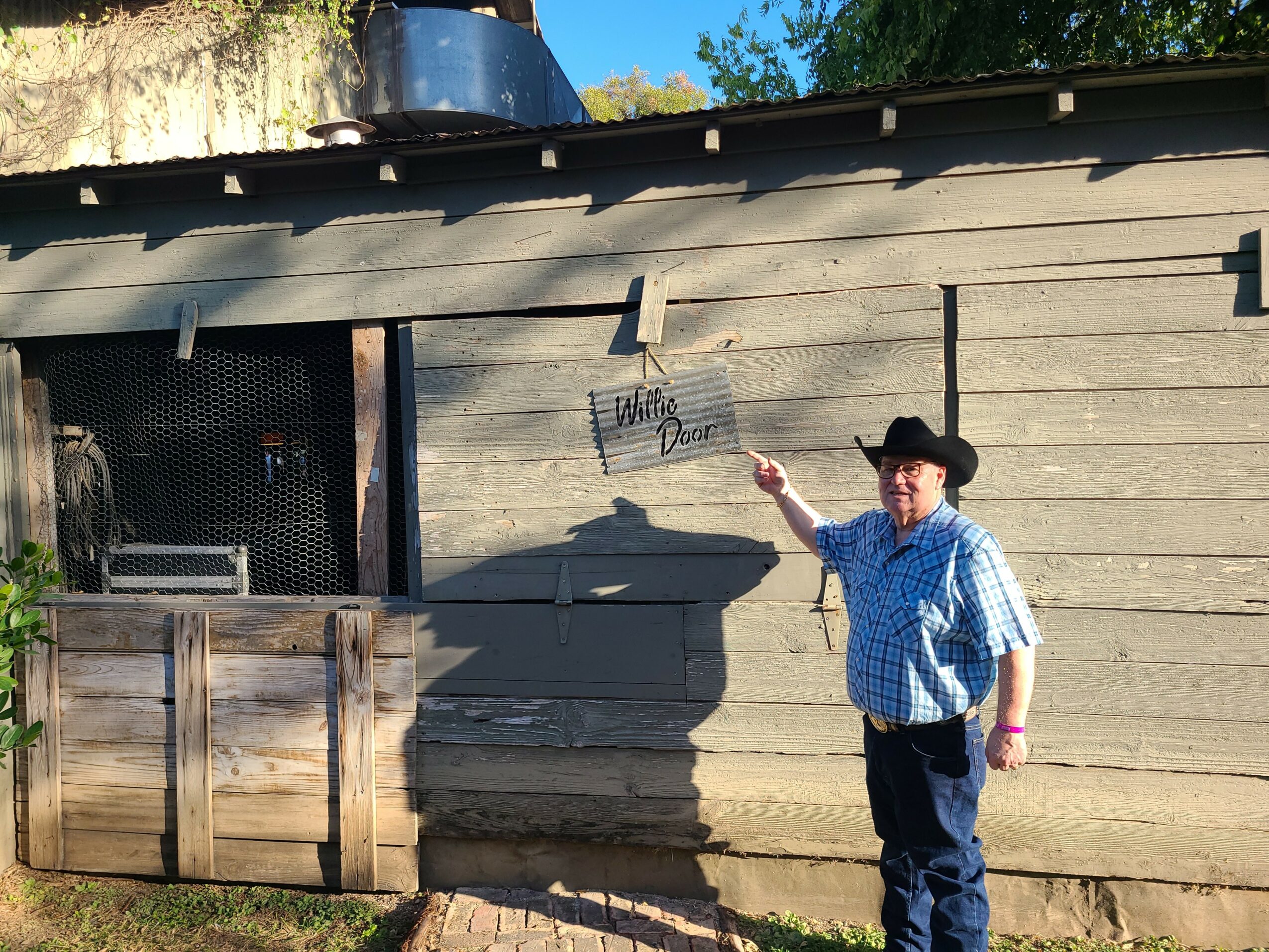Child wearing a cowboy hat and plaid shirt pointing at a wooden wall outside.