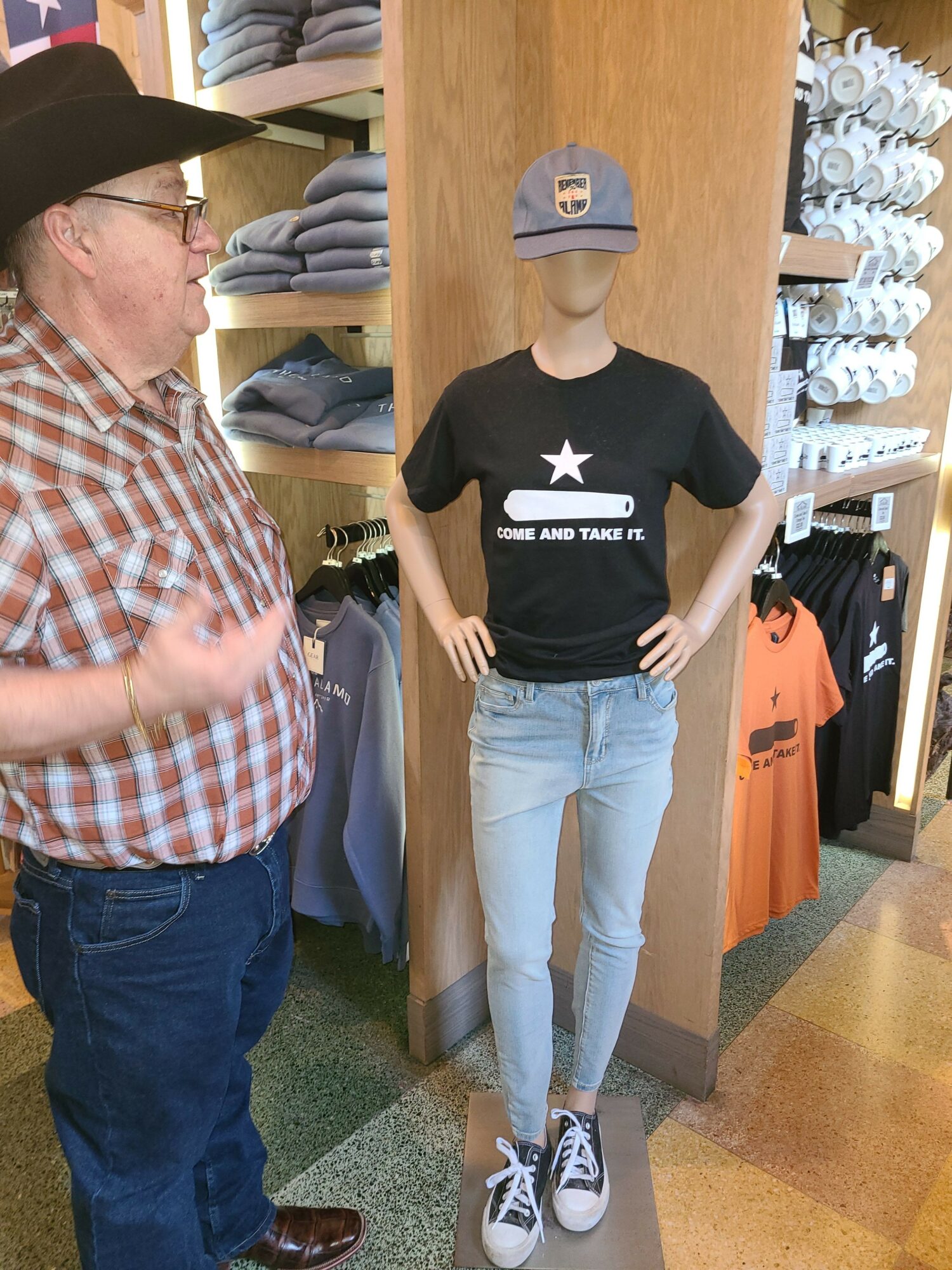 Man in plaid shirt and glasses talking to a mannequin wearing a black t-shirt, jeans, and a cap in a store.