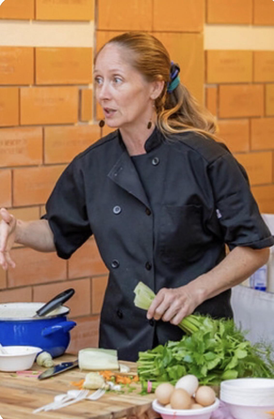 Woman in black chef coat gesturing with her right hand, standing in front of a brick wall, with eggs and greens on the table.