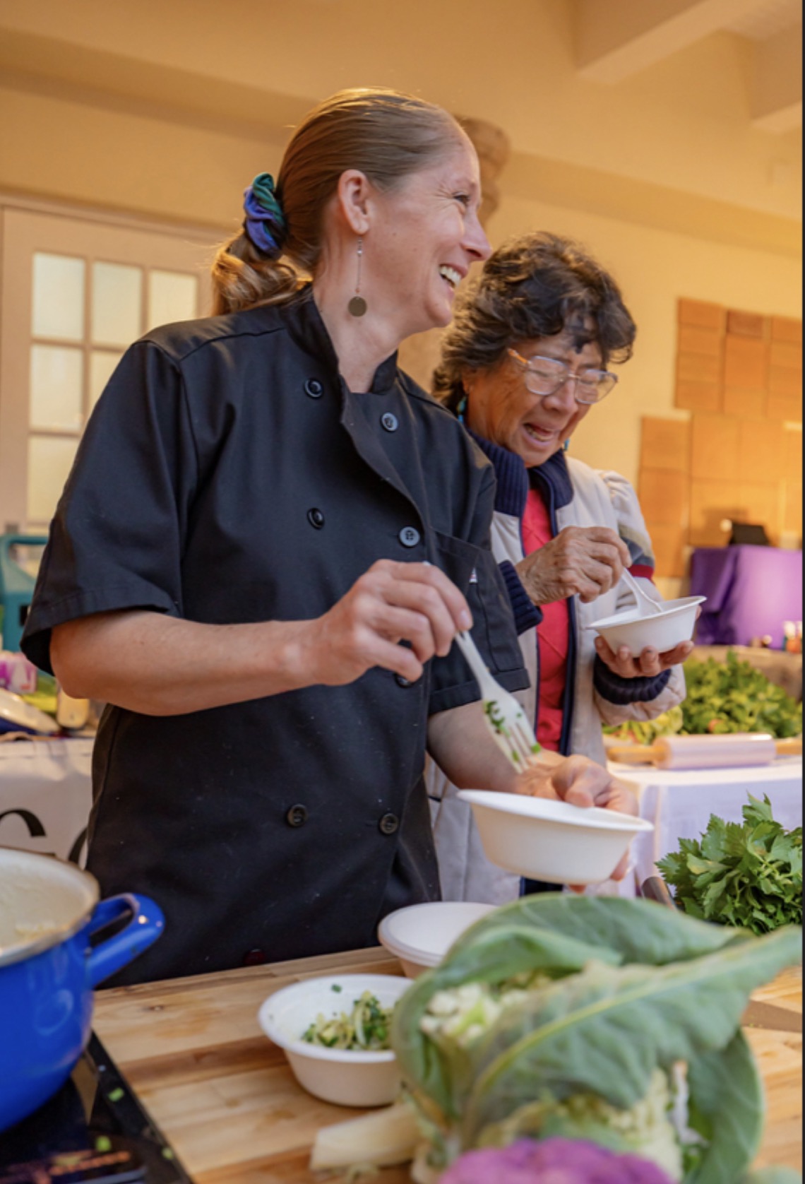 Two women cooking and smiling in a kitchen with vegetables and bowls on the counter.