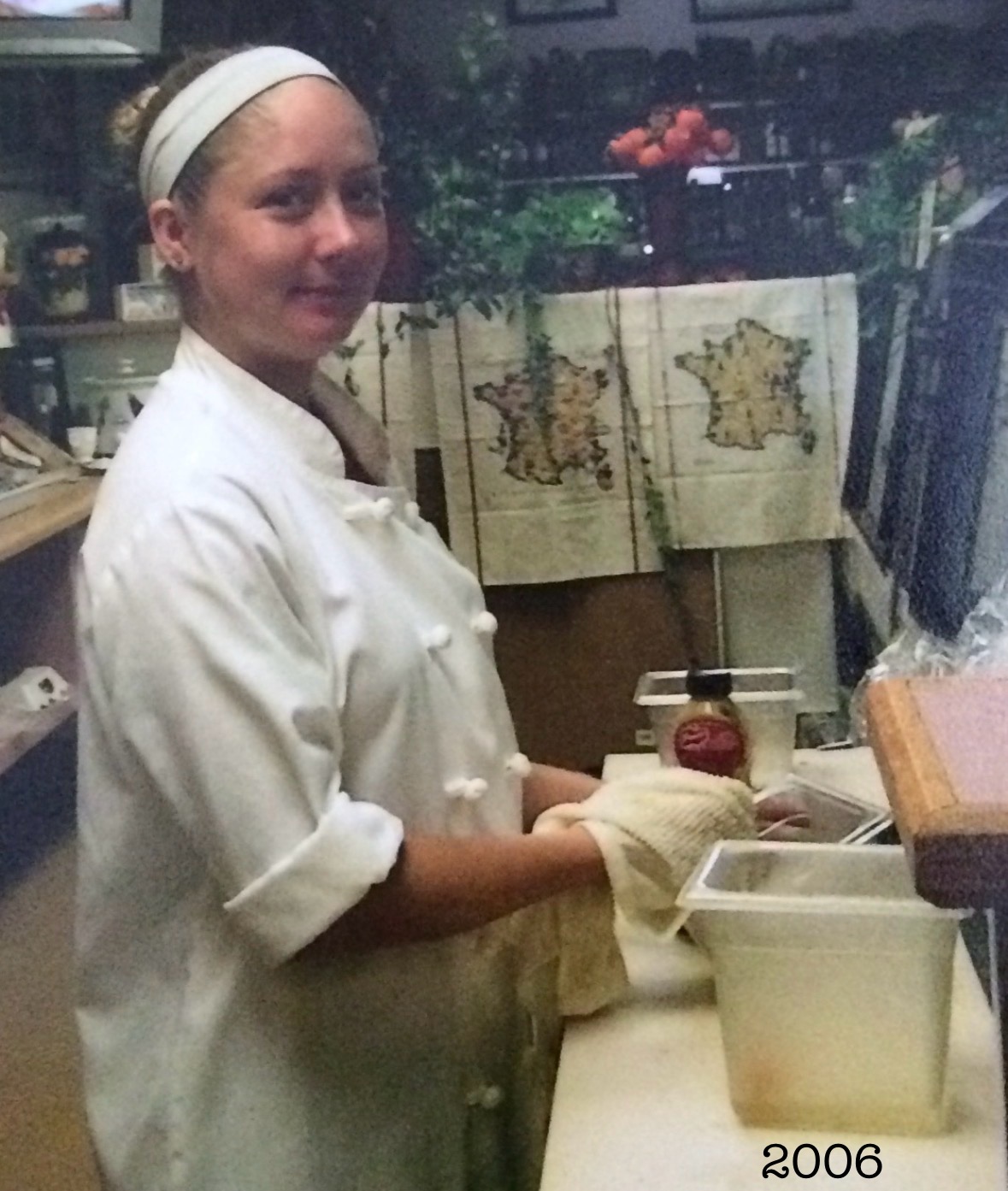 Woman in chef's uniform and gloves standing in kitchen with shelves and containers behind her.