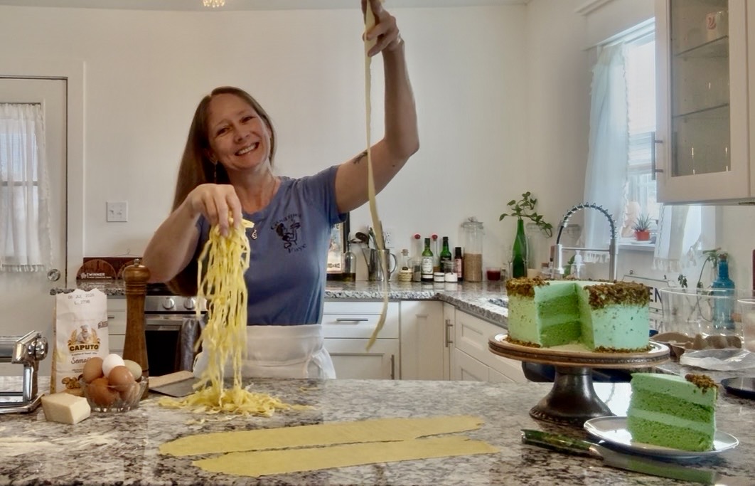 Woman in kitchen holding pasta with one hand, smiling, with a cake on a stand nearby.