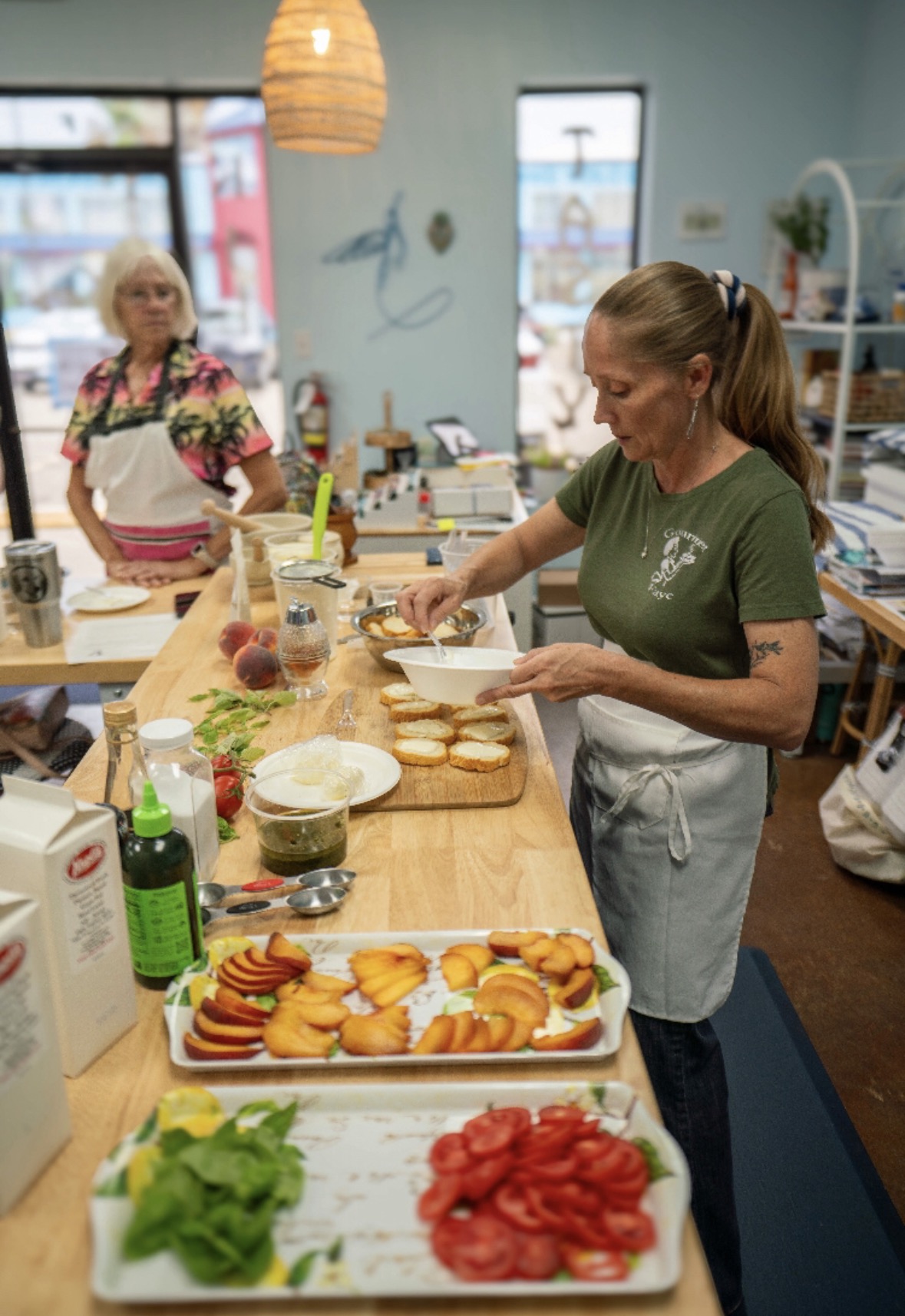 Two women preparing food in a kitchen with a wooden counter, various ingredients, and a window in the background.
