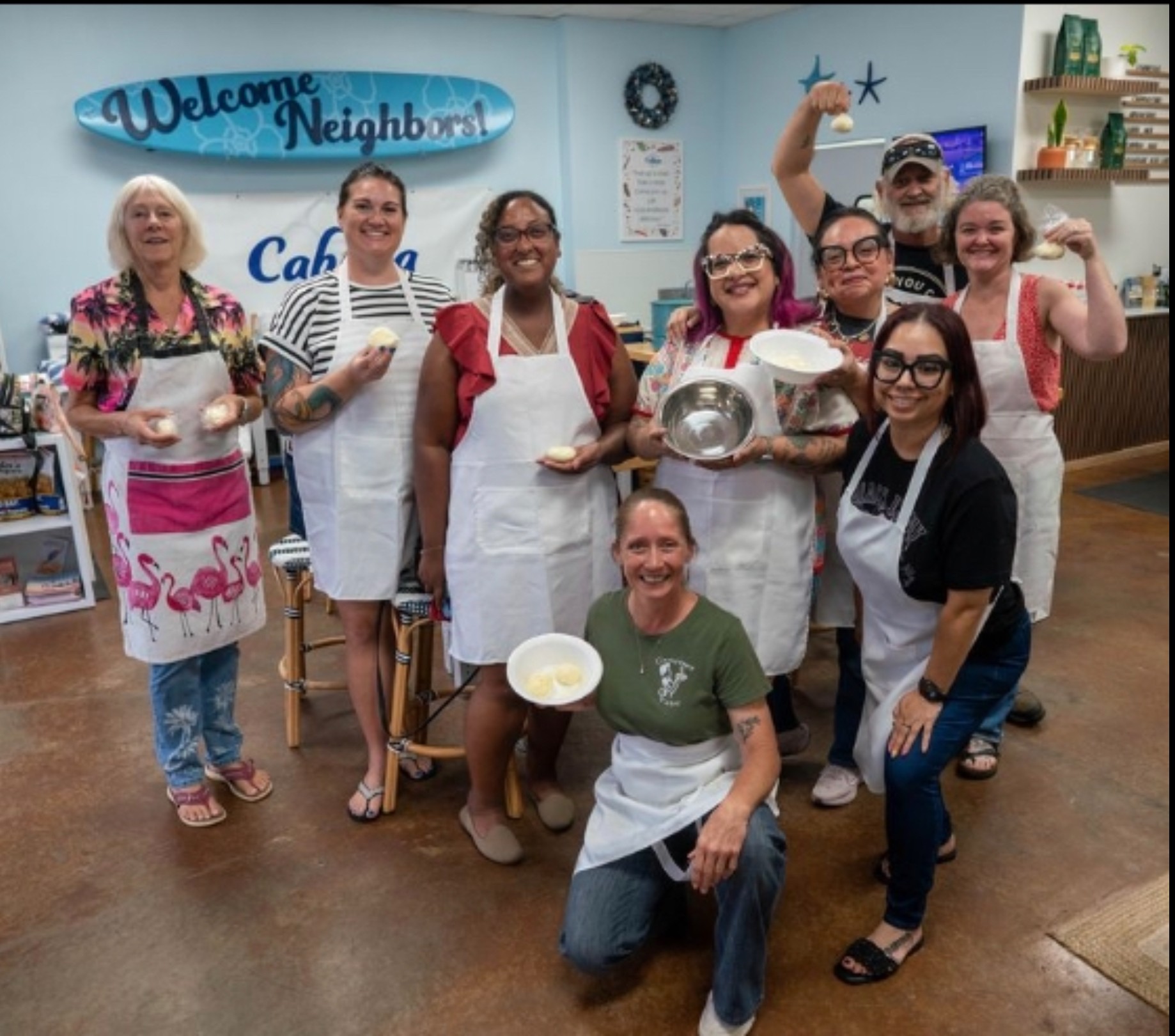Group of nine people smiling, holding bowls and utensils, in a room with a welcome sign and shelves.