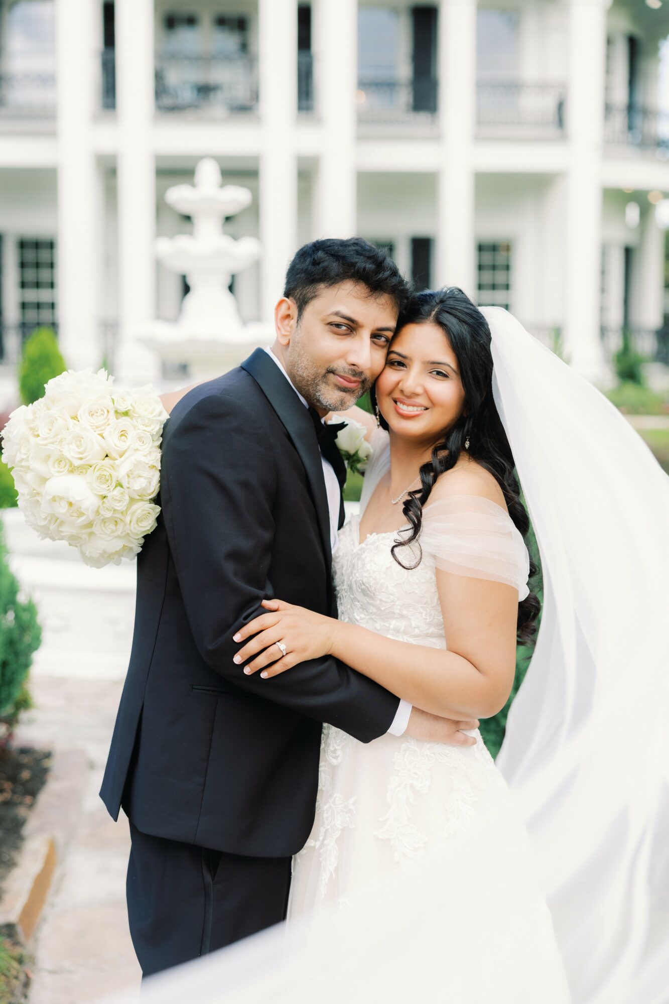 Bride and groom smiling, embracing outdoors with a white building and fountain in background.