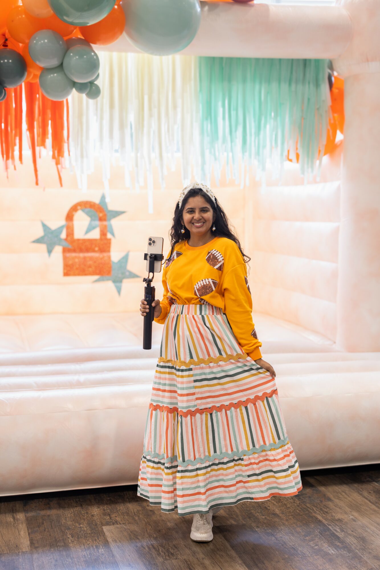 Woman in yellow top and striped skirt standing in front of colorful backdrop with balloons and stars, smiling.