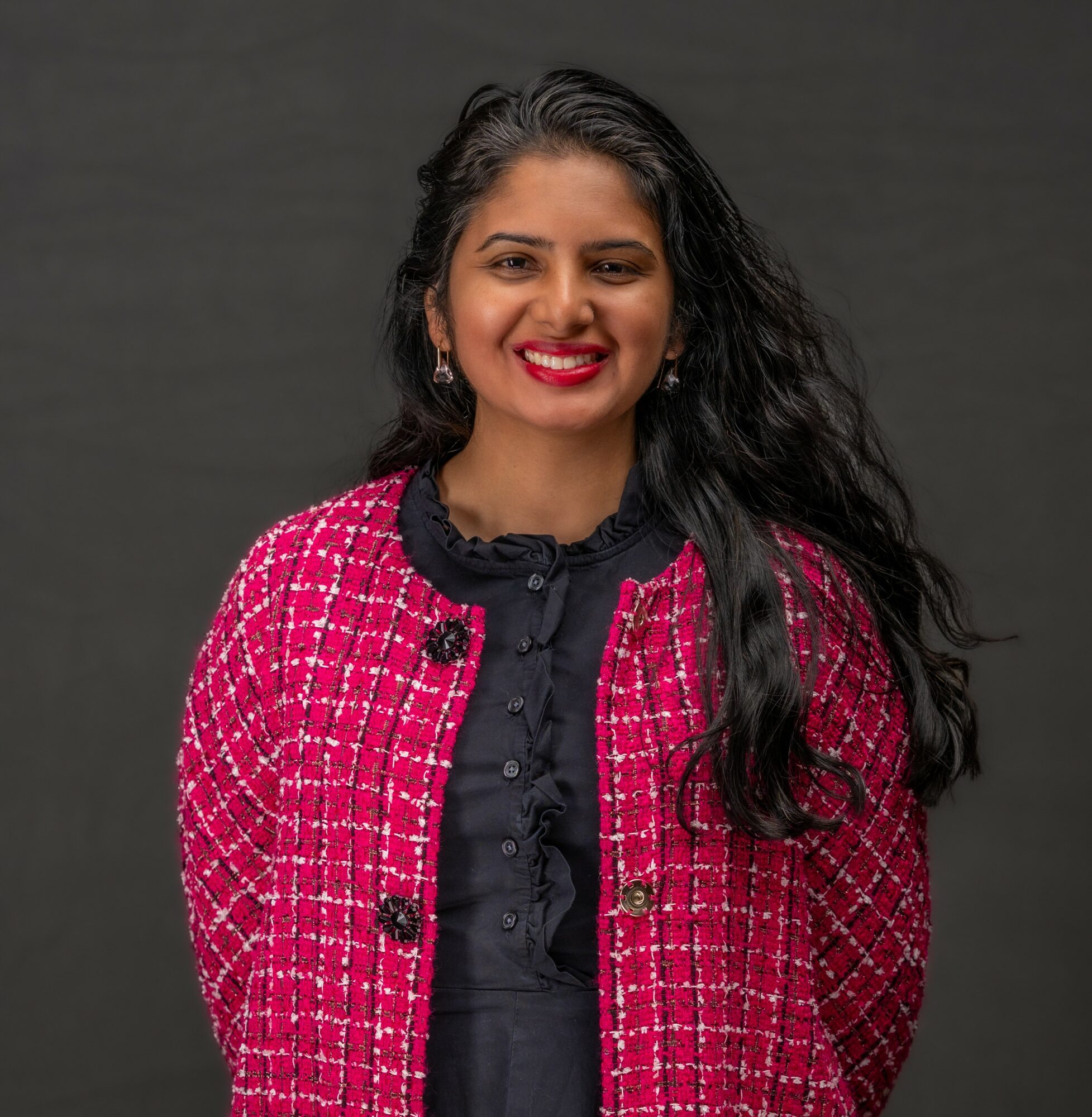 Woman with long dark hair, wearing a pink and black patterned jacket and black blouse, smiling against a dark background.