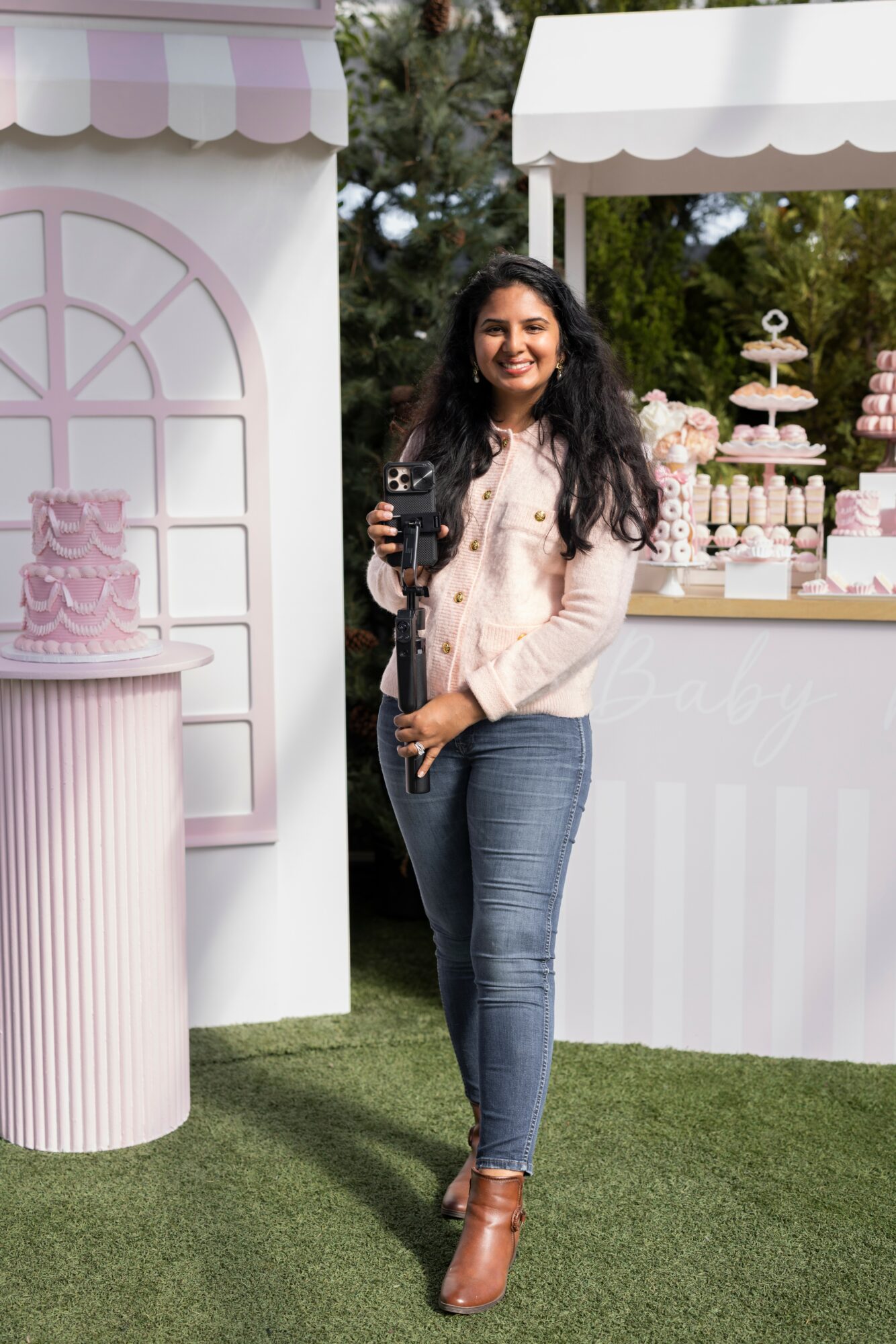 Woman with long dark hair holding a glass, standing outdoors near a dessert stand and decorative backdrop.