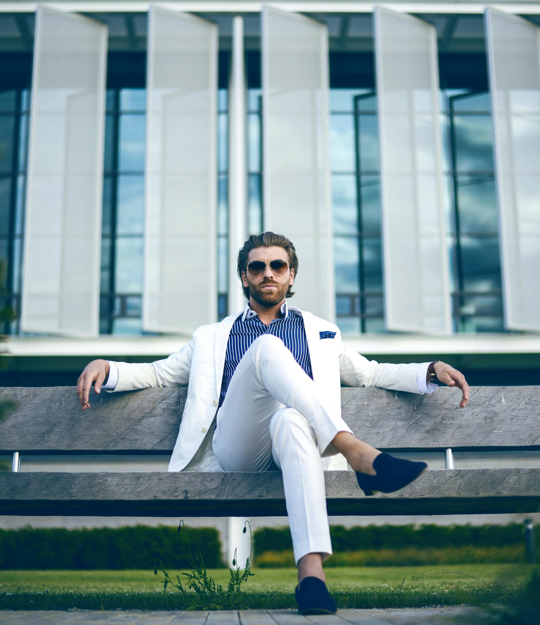 Man with sunglasses in white suit sitting on a bench outside a modern glass building.