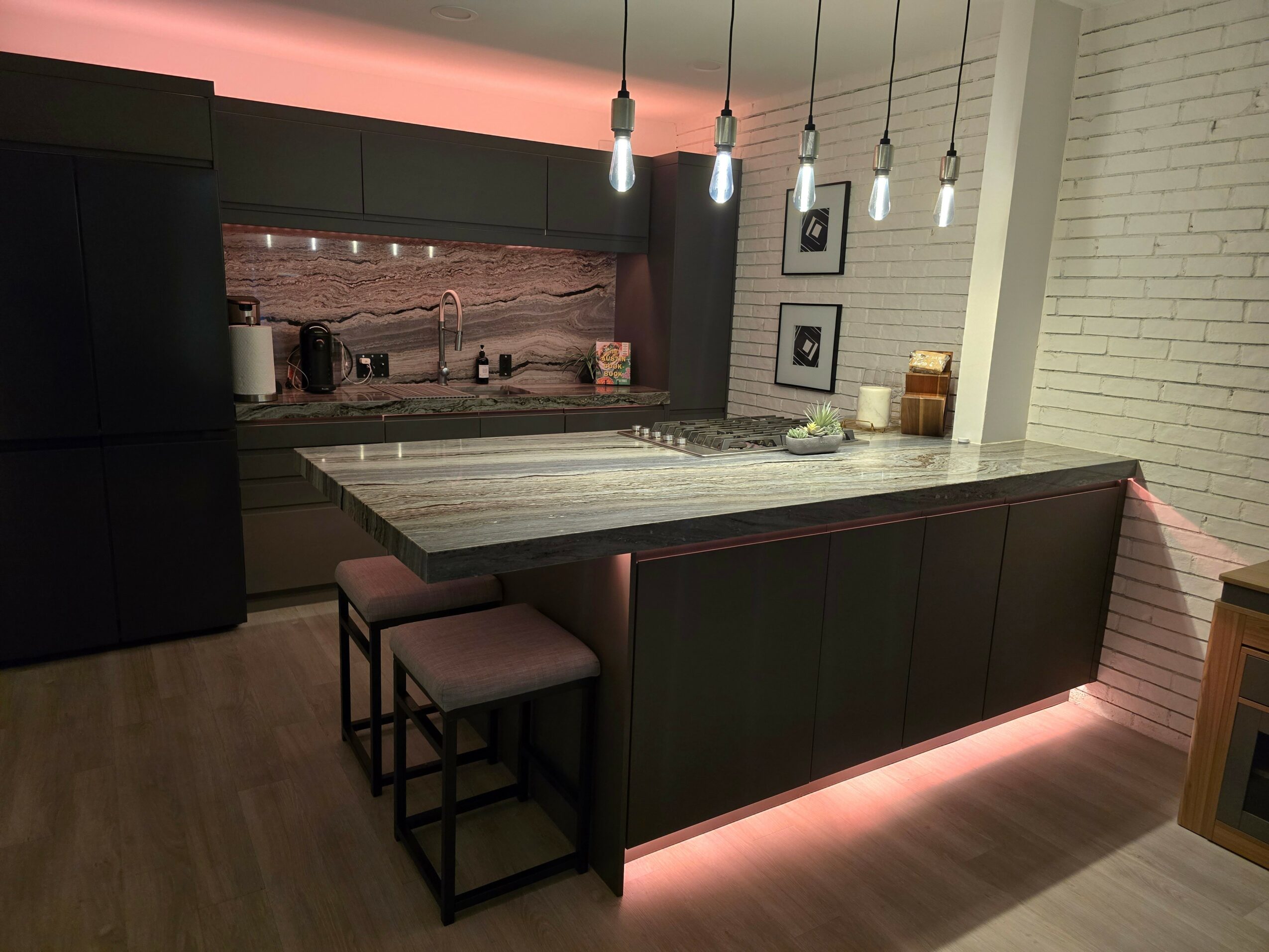 Kitchen with a marble island, hanging pendant lights, black cabinets, and a white brick wall, illuminated by pink accent lighting.