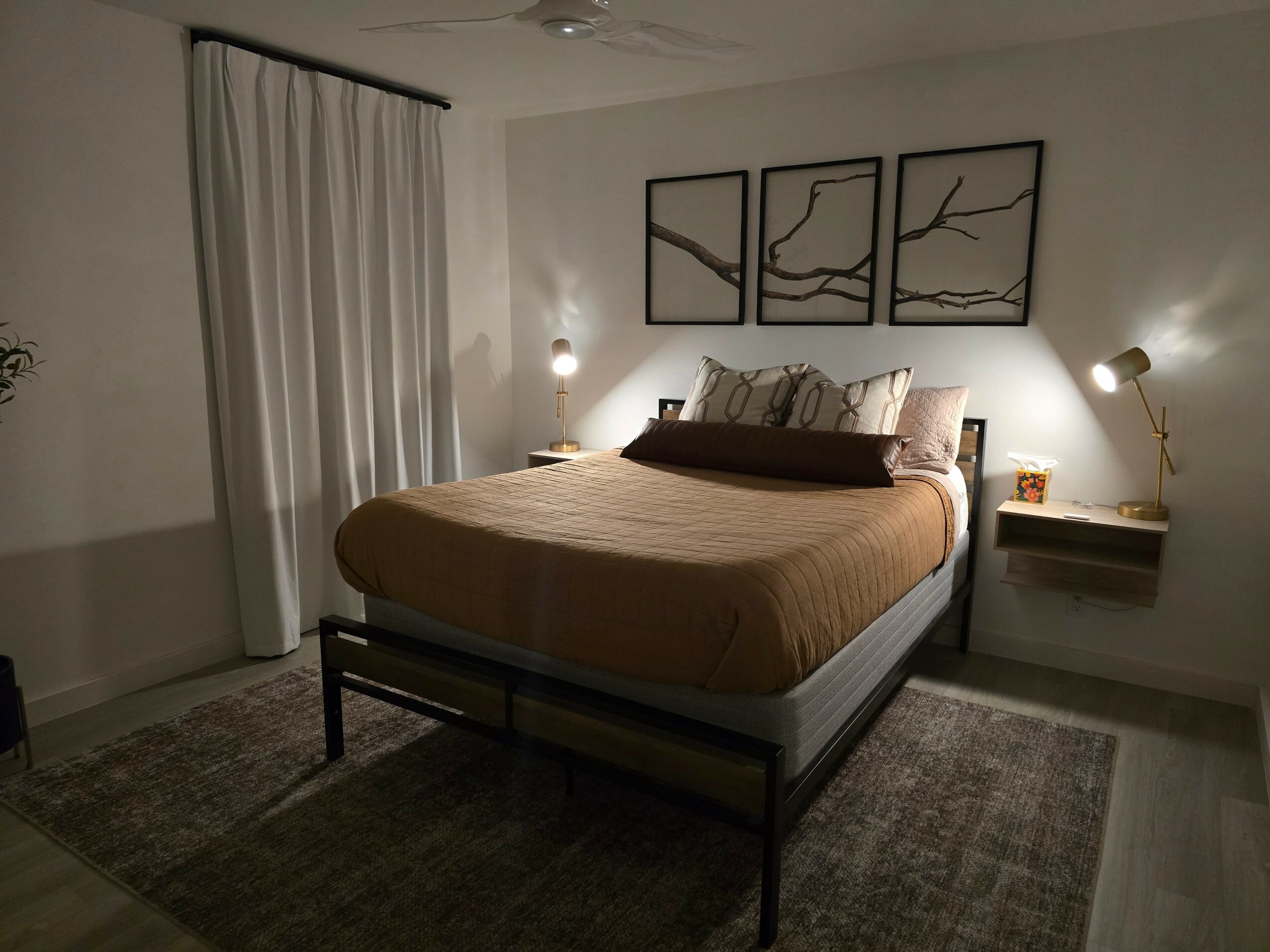 Bedroom with bed, wall art, two wall-mounted lamps, and a side table with a lamp, curtains, and a rug.