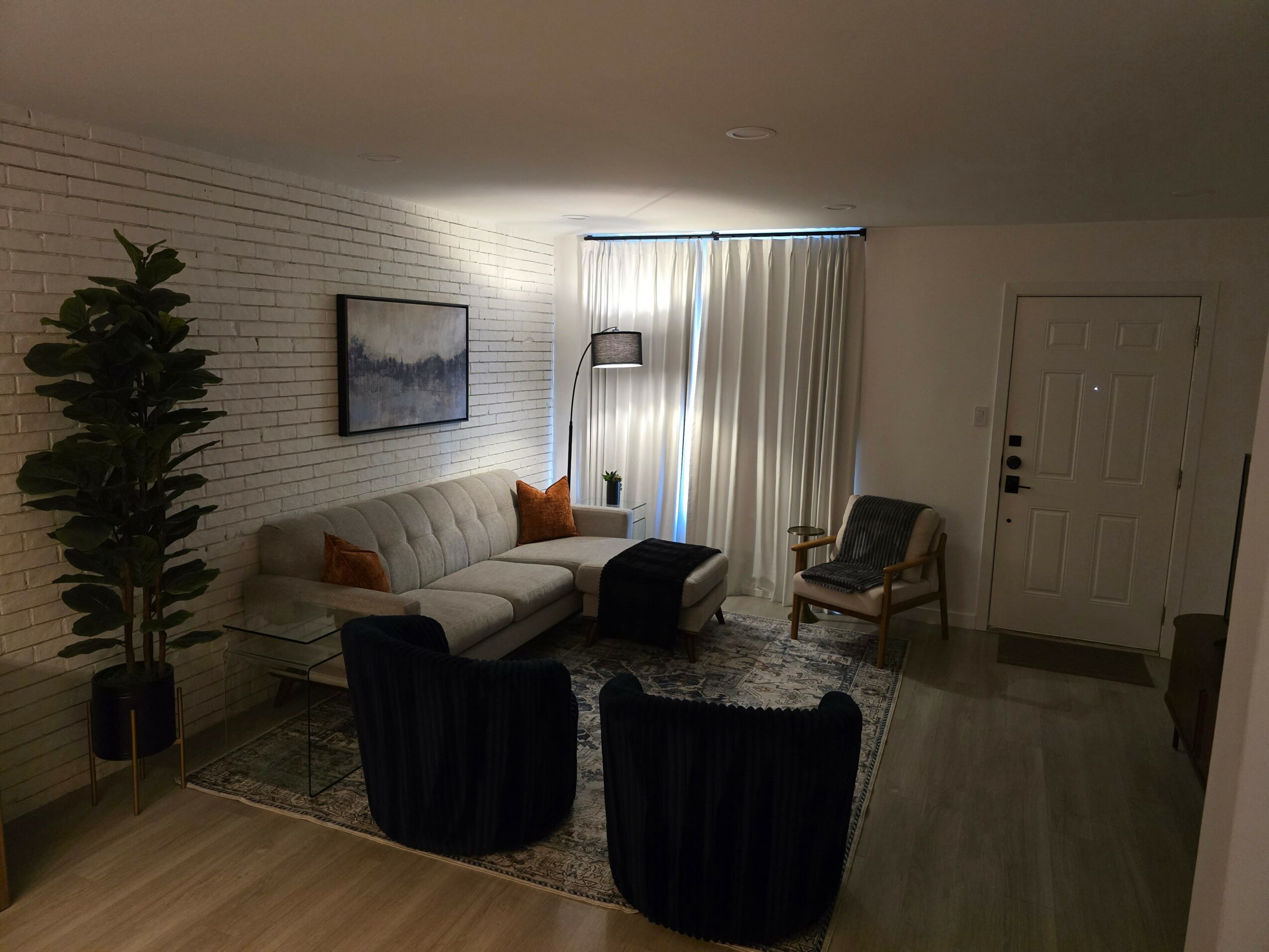 Living room with white brick wall, sofa, two armchairs, tall plant, and window with curtains.