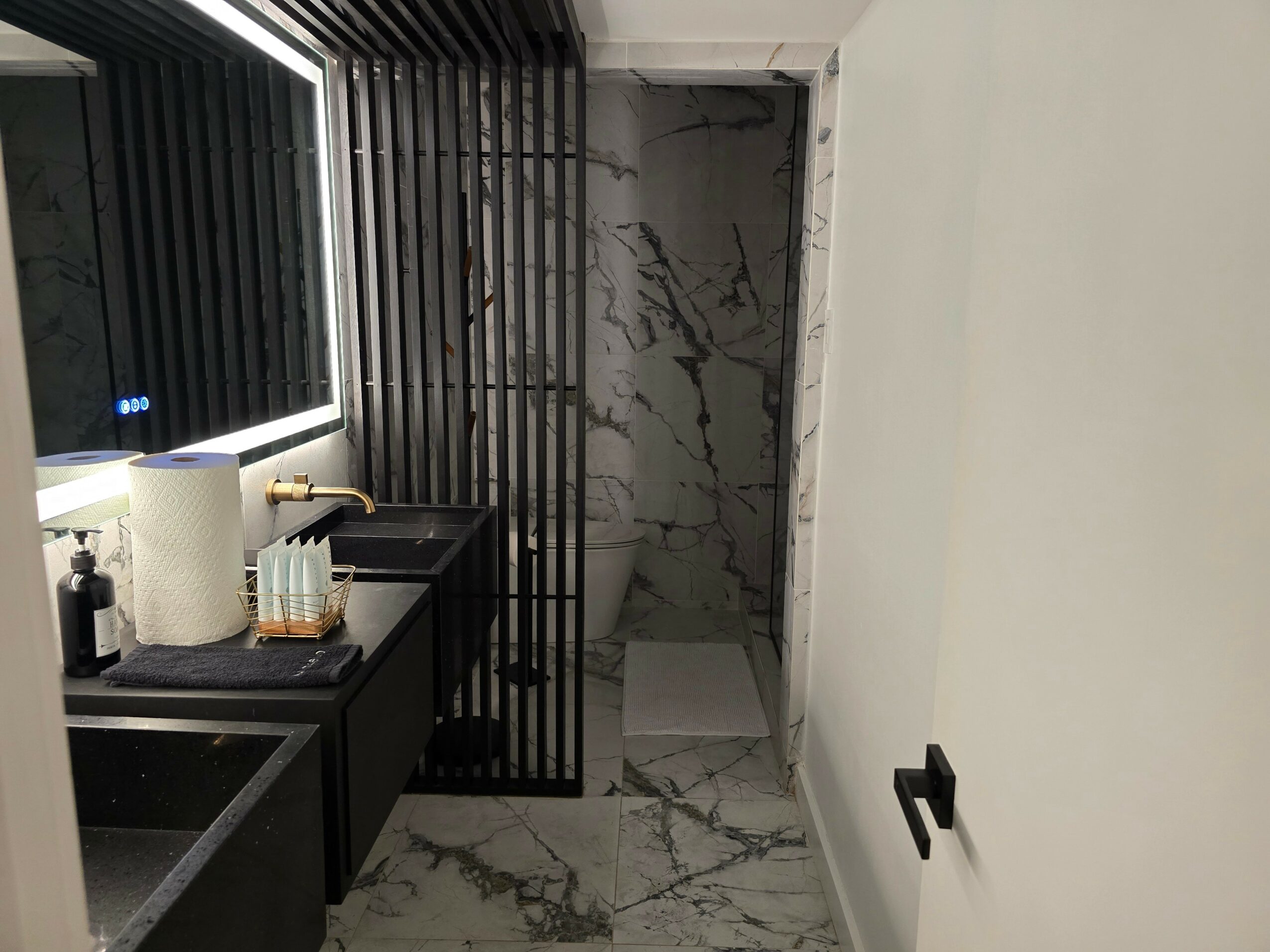 Bathroom with black and white marble flooring, a black countertop with paper towels, and a shower area with marble walls.