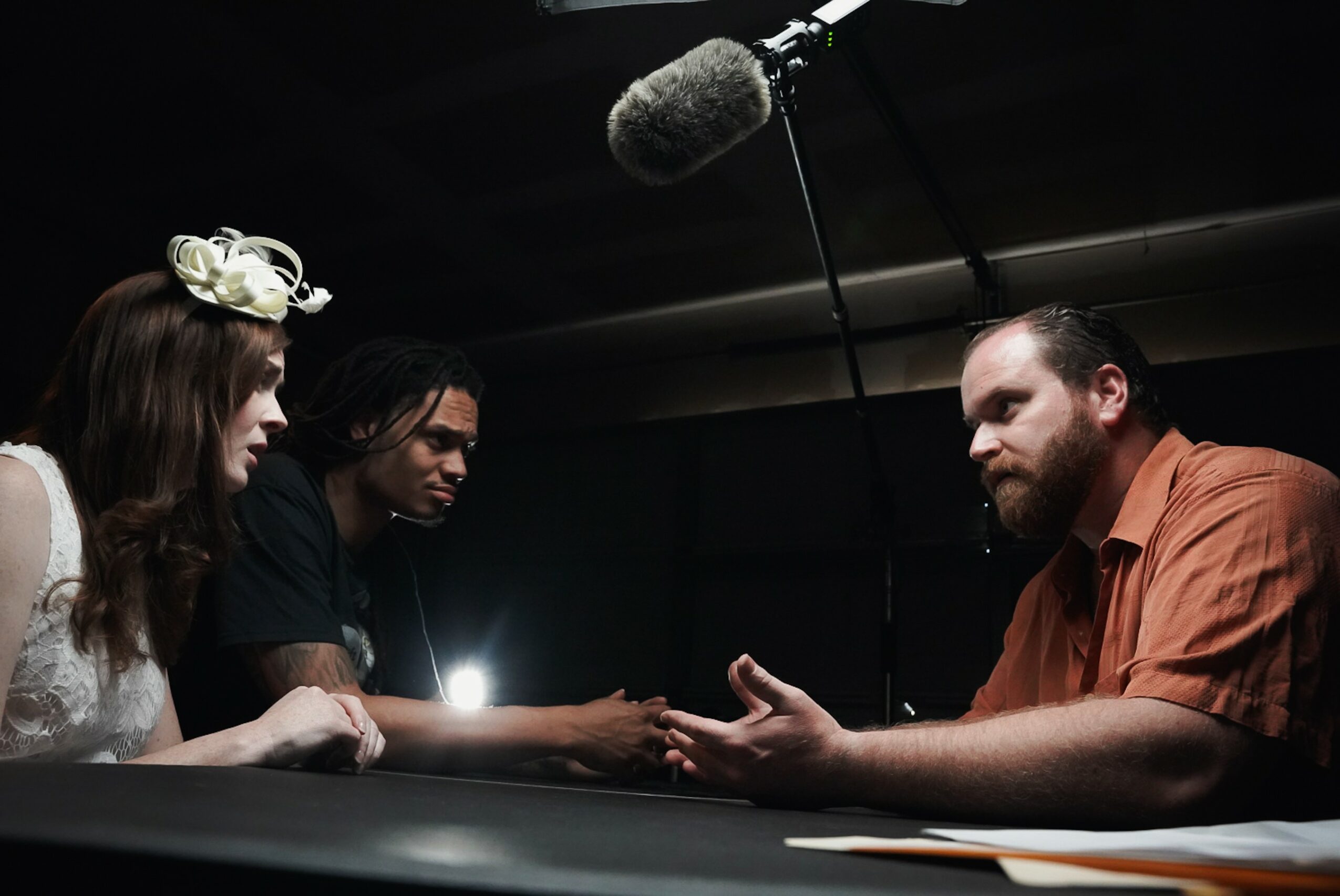 Two men and a woman sit at a table, with a microphone hanging above, engaging in a conversation or interview.
