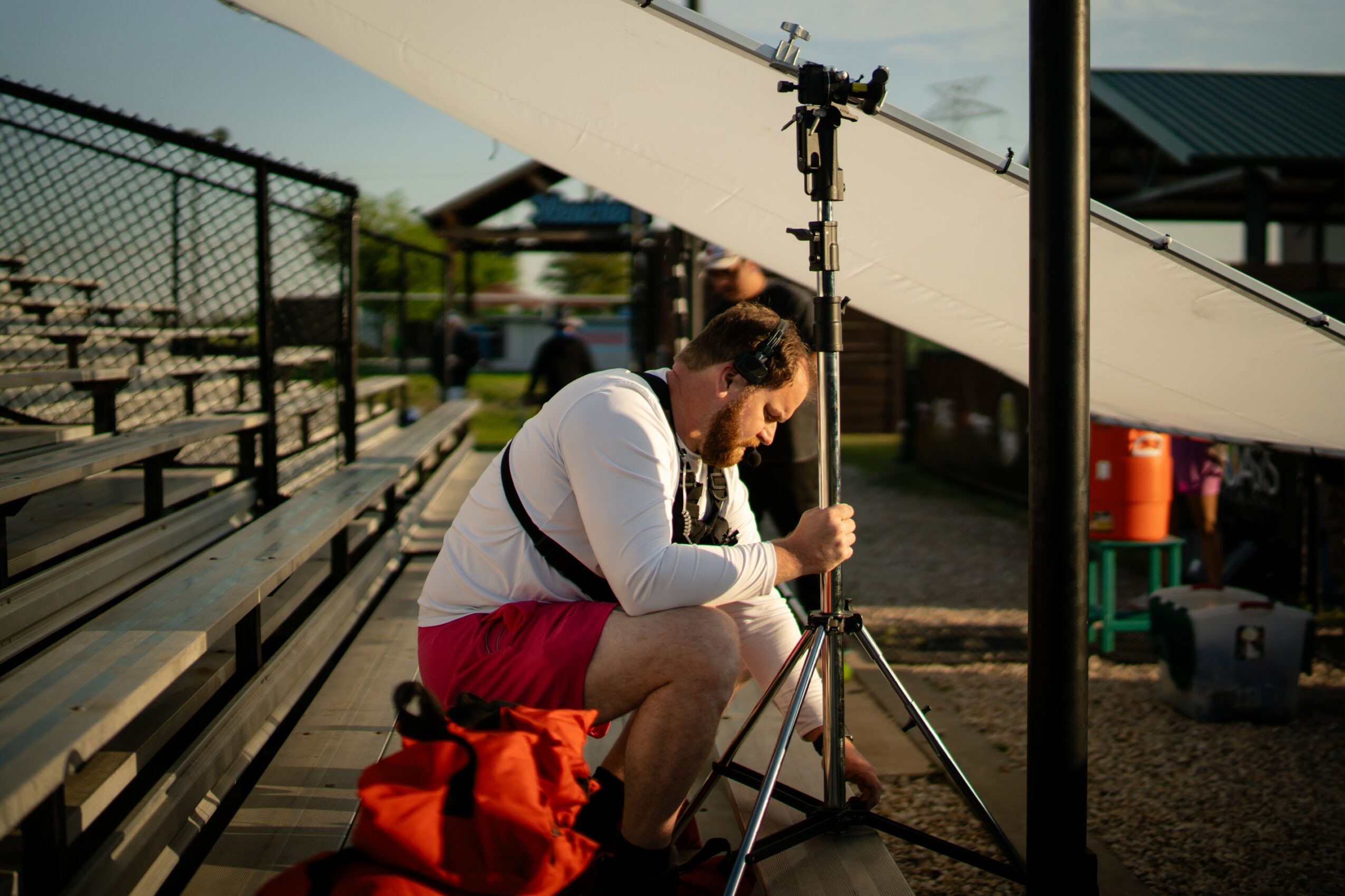 Person adjusting camera on tripod outdoors near bleachers and a chain-link fence.