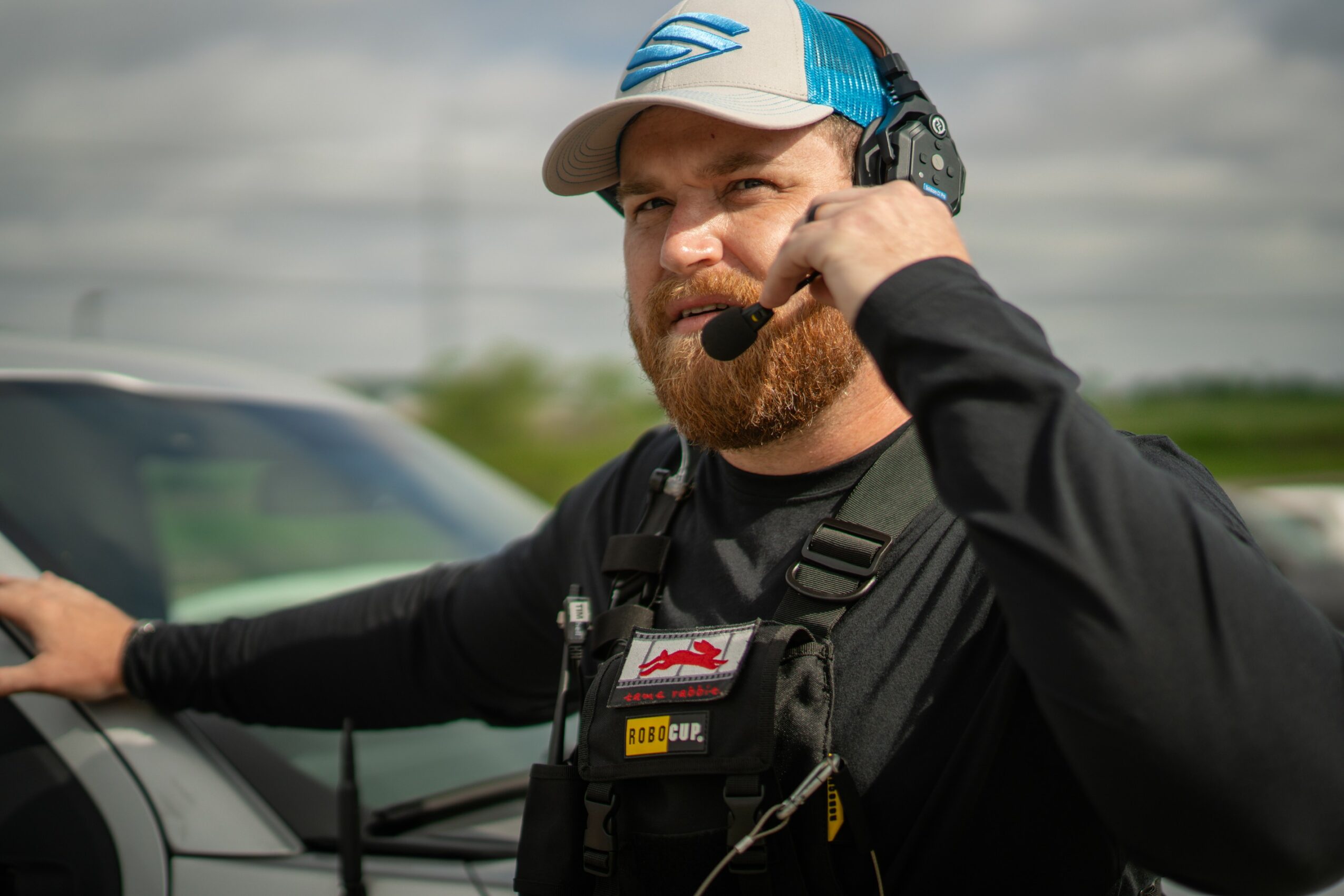 Man with a beard wearing a cap and headset, talking on a communication device outdoors near a vehicle.