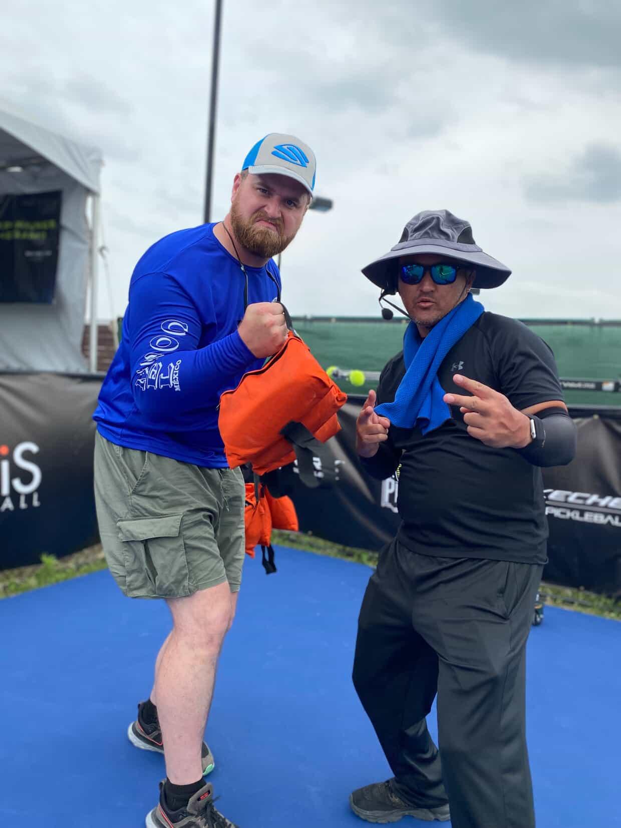 Two people standing outdoors on a blue surface, one holding a small orange object, the other making a peace sign.