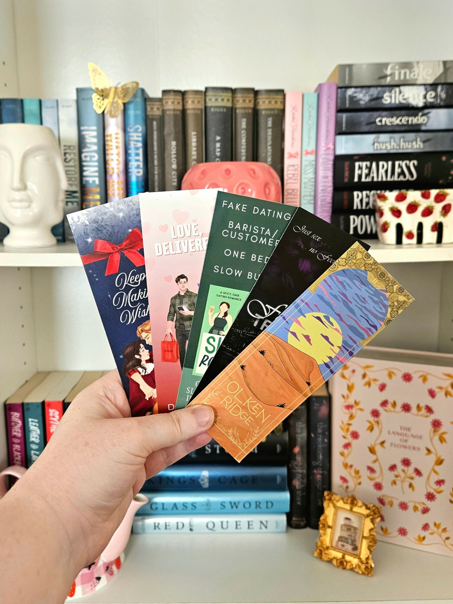 Hand holding five colorful bookmarks in front of a bookshelf with books and decorative items.