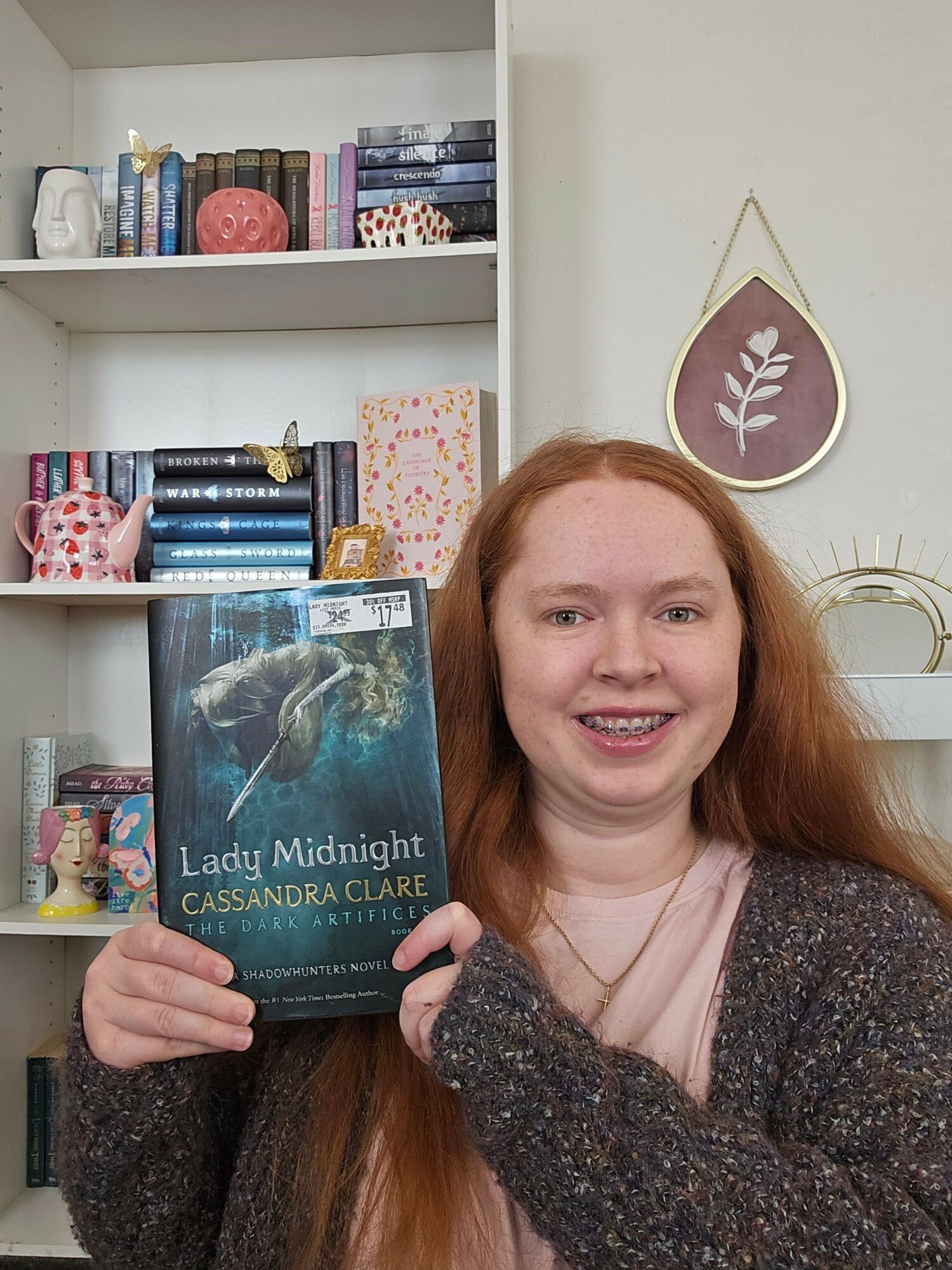 Young woman with long red hair holding a book titled 'Lady Midnight' in a room with shelves and wall decor.