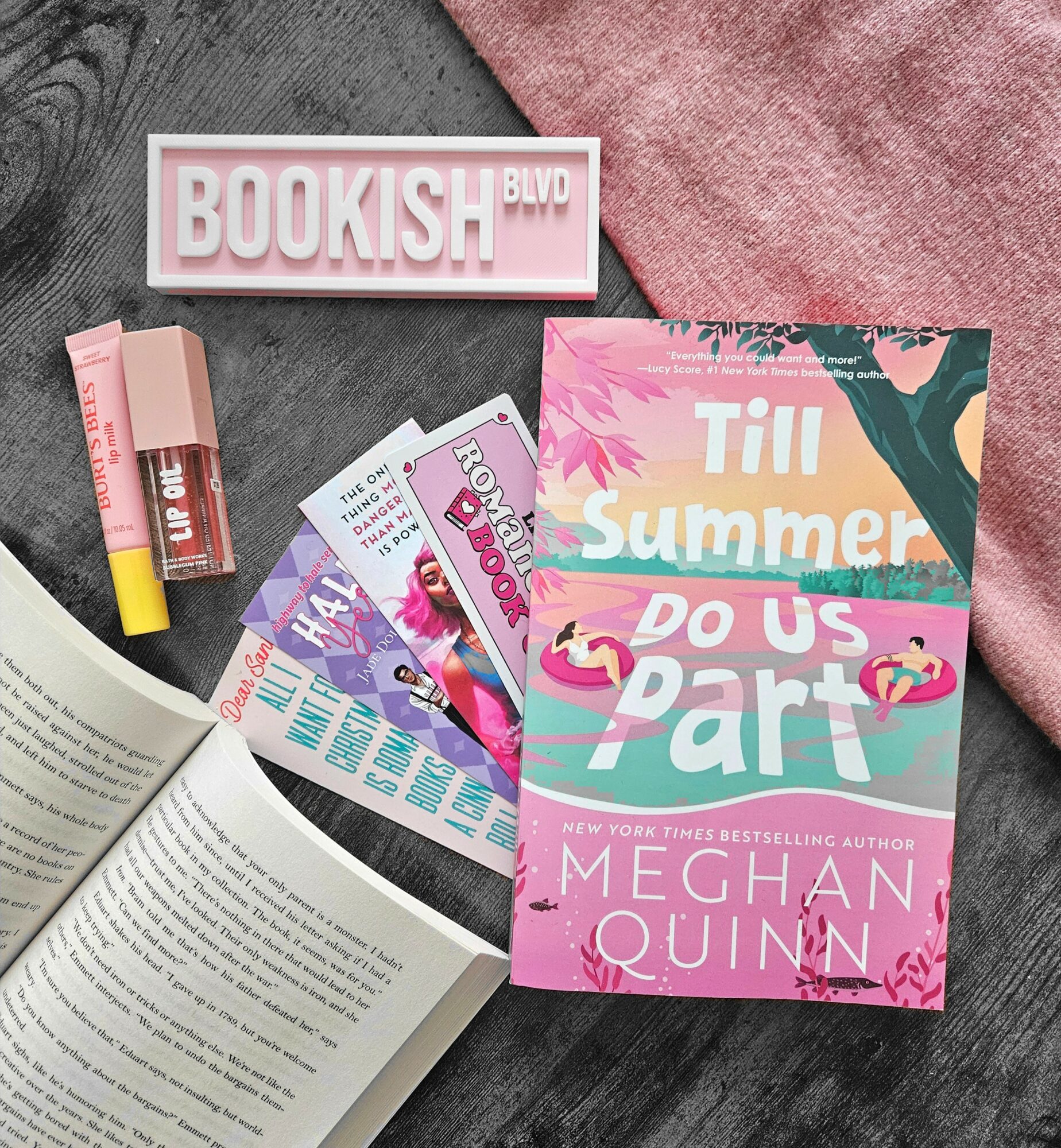 Open book, pink book titled 'Till Summer Do Us Part,' pink and white sign reading 'BOOKISH BLVD,' lip gloss, and pink fabric.