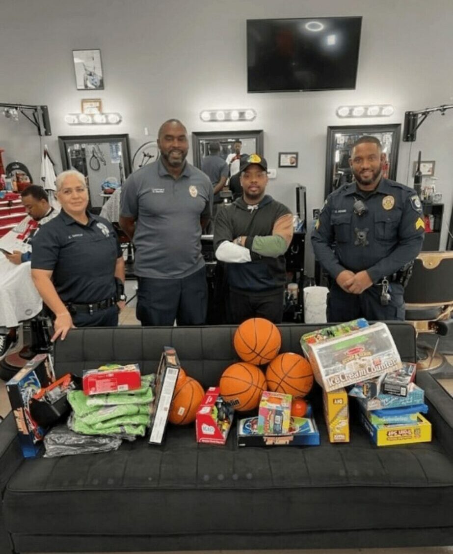 Four people standing behind a table with basketballs, toys, and supplies in a room with a TV and shelves.