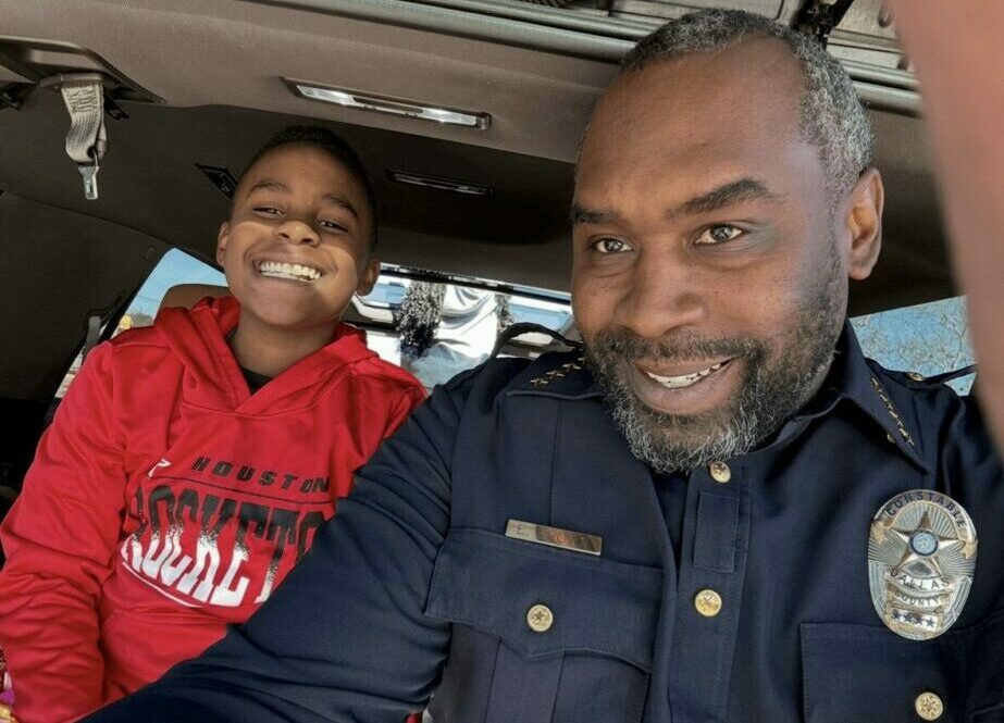 Man and child smiling inside a vehicle, man wearing a police uniform, child in red hoodie.