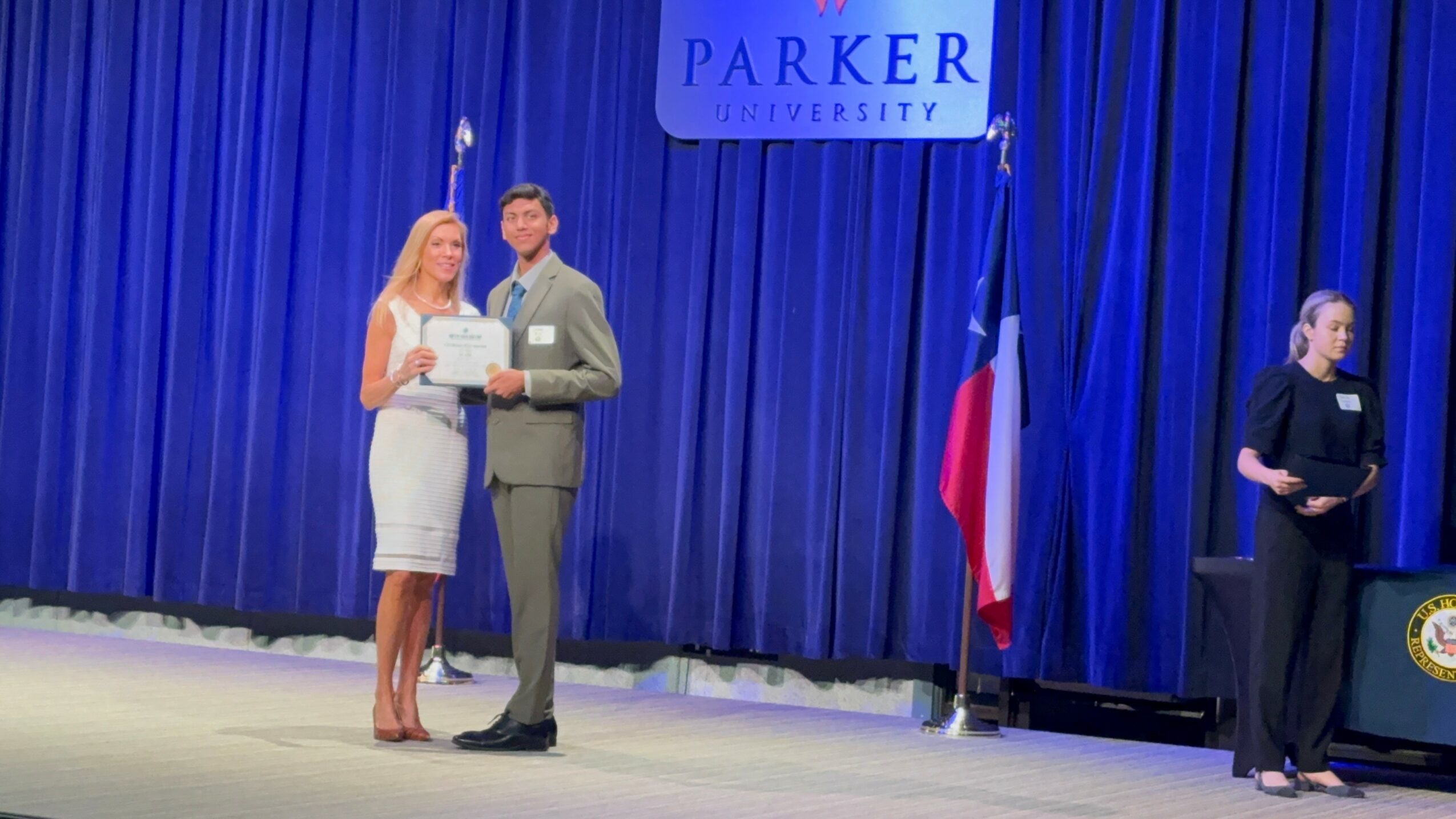 Two people standing on stage with a blue curtain background, a sign, and a flag, holding a certificate or award.