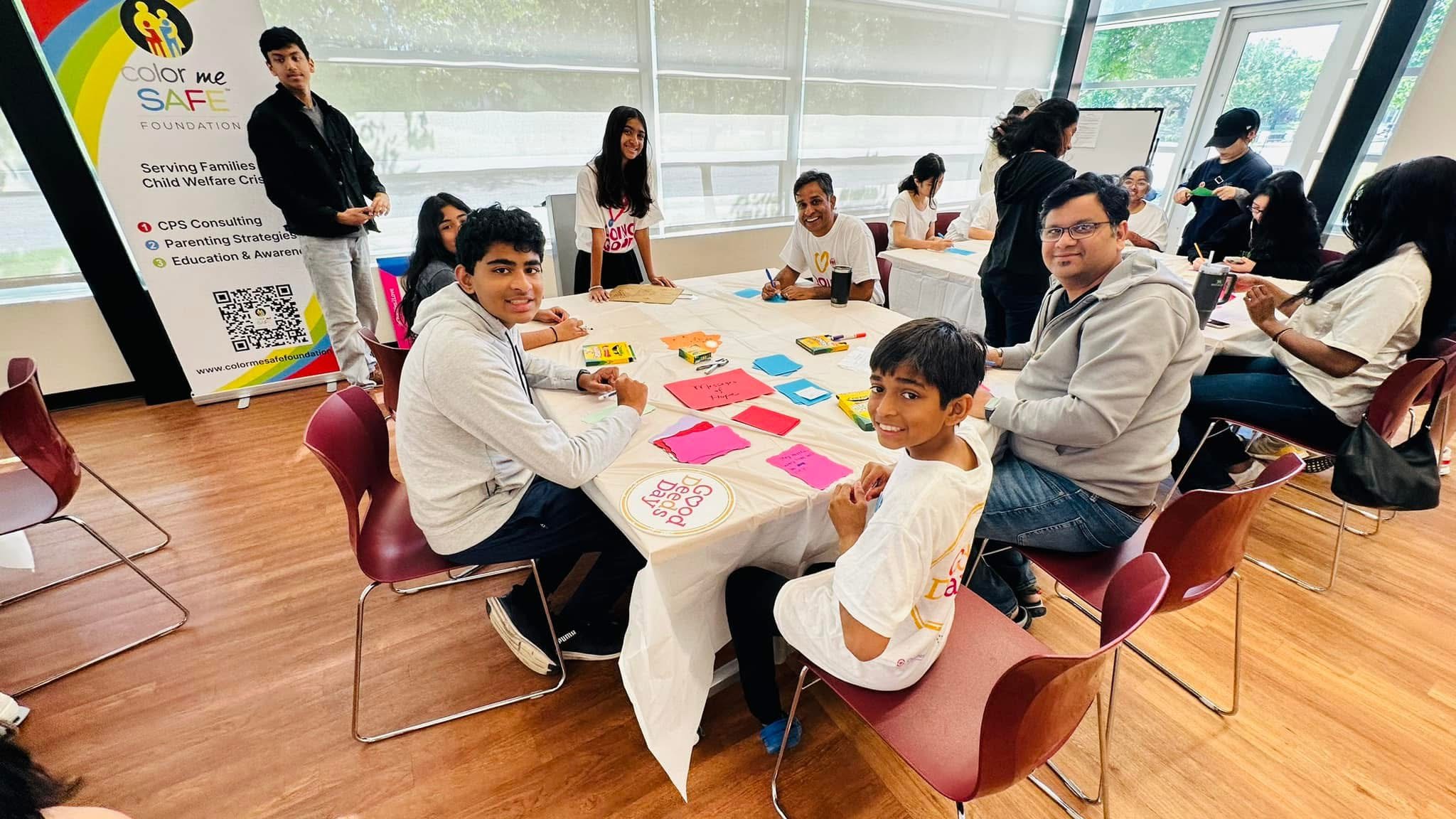 Group of children and adults sitting around a table with colorful paper and markers in a bright room.
