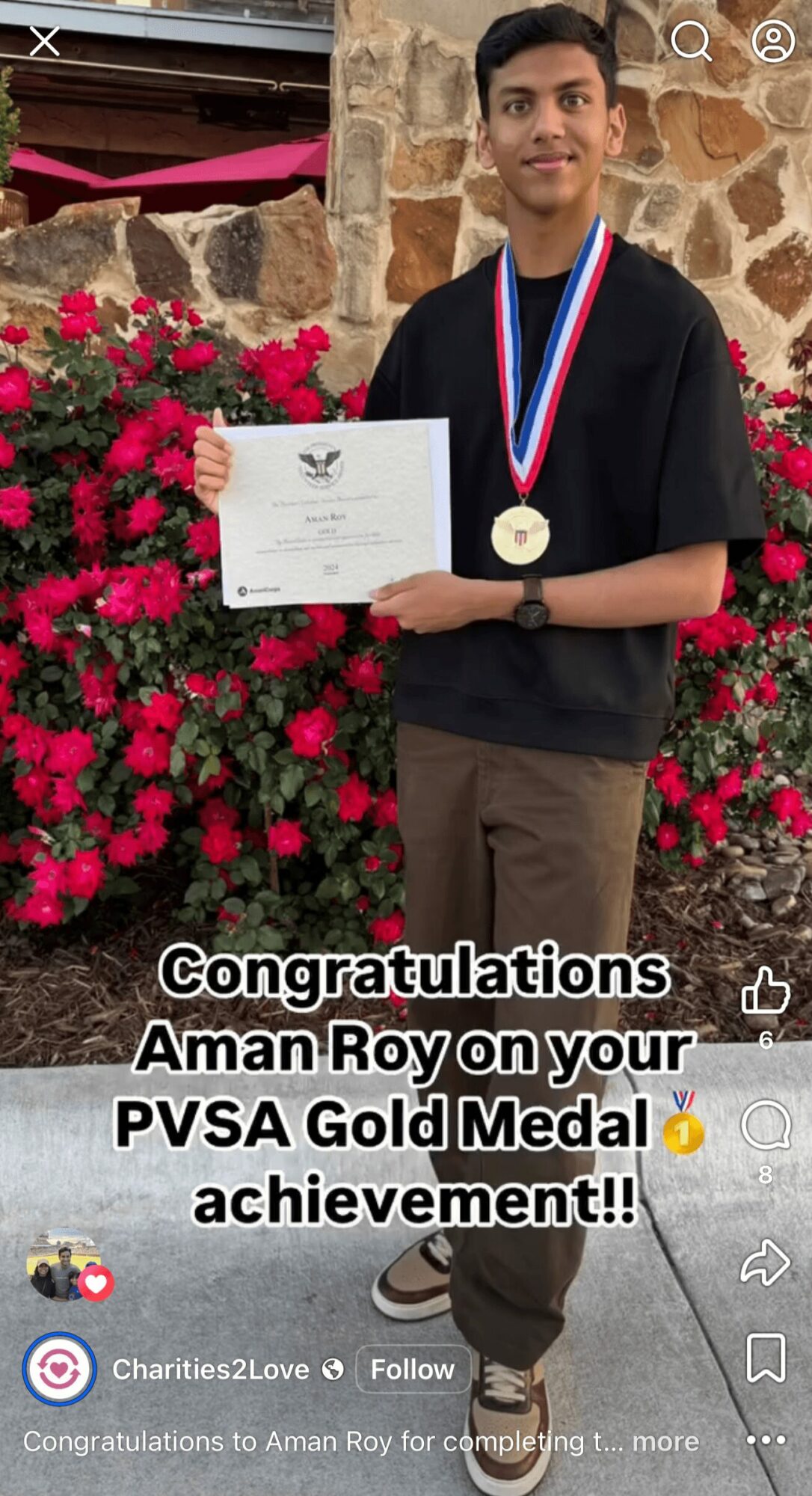 Young man holding a certificate and wearing a gold medal, standing in front of pink flowers and stone wall.