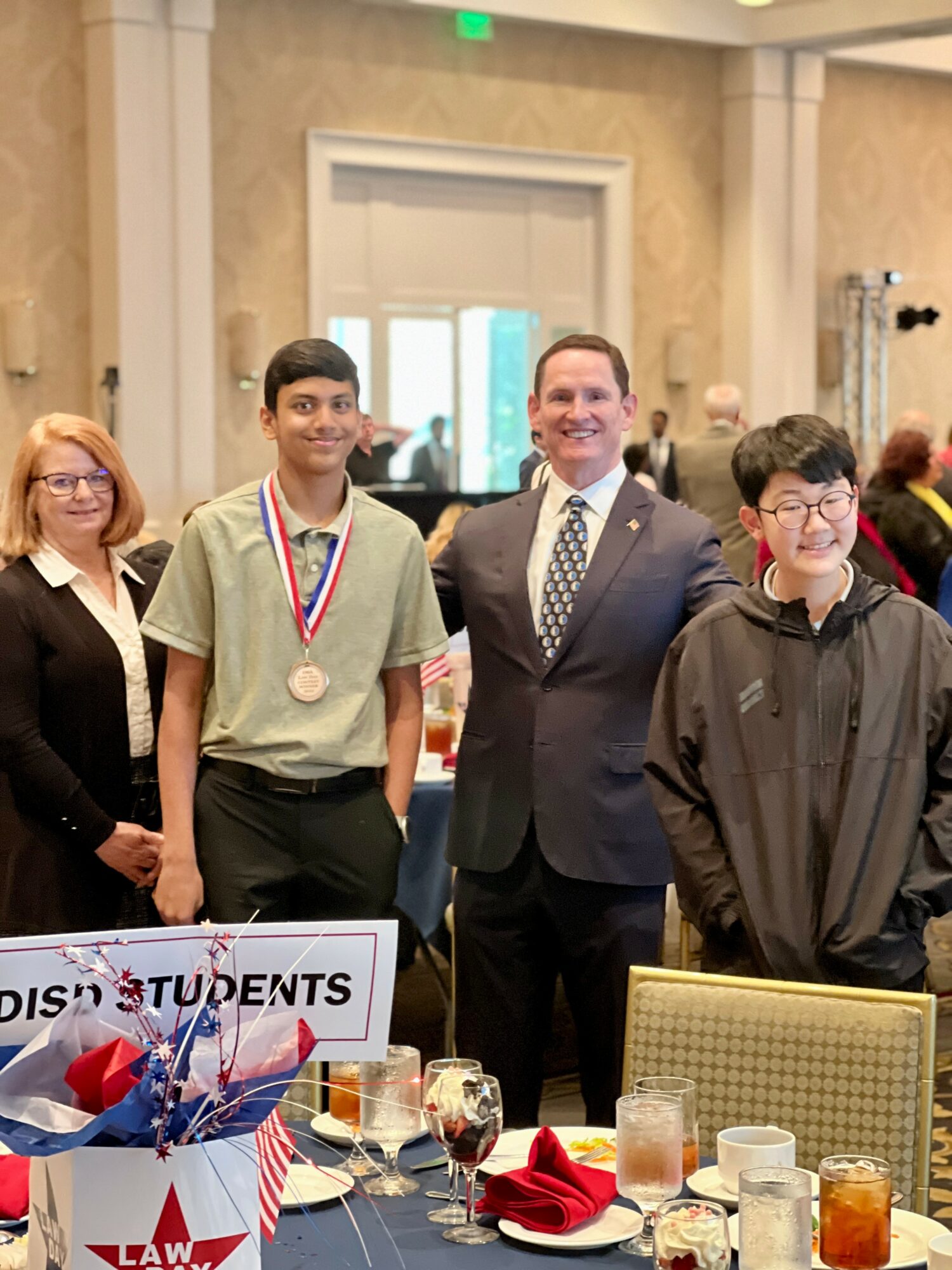 Group of four people standing indoors, one person wearing a medal, table with drinks and a sign in front.