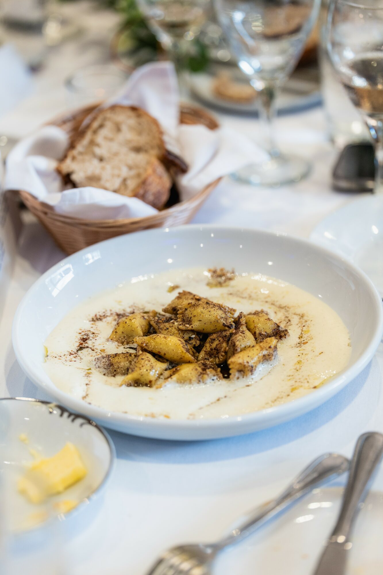Creamy dish with chunks of meat and herbs in a white bowl on a table with bread and wine glasses.