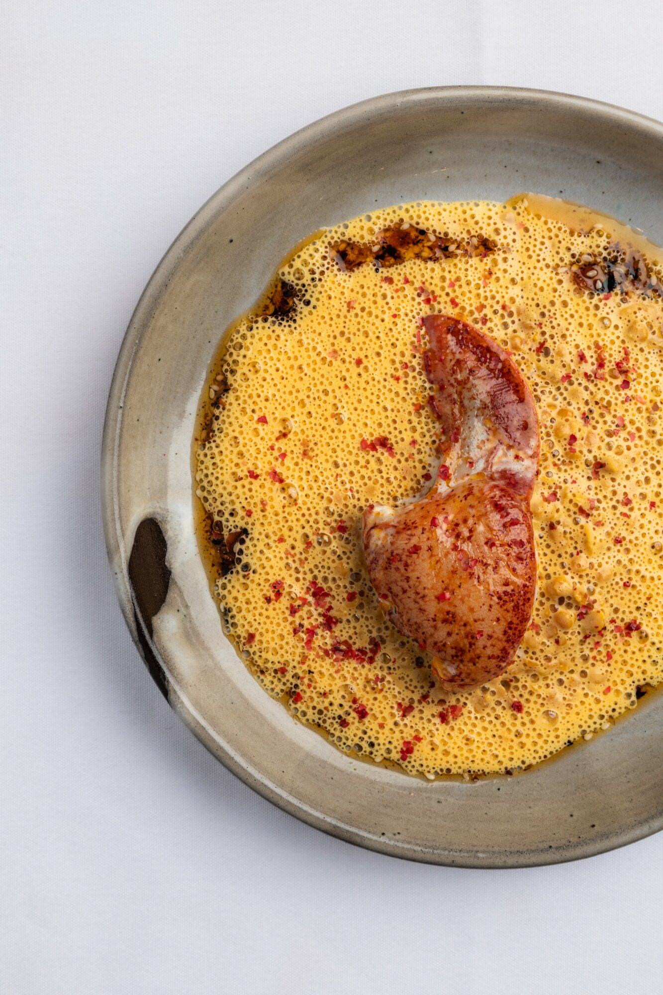 A bowl with a piece of meat submerged in a yellow, frothy sauce with red specks.