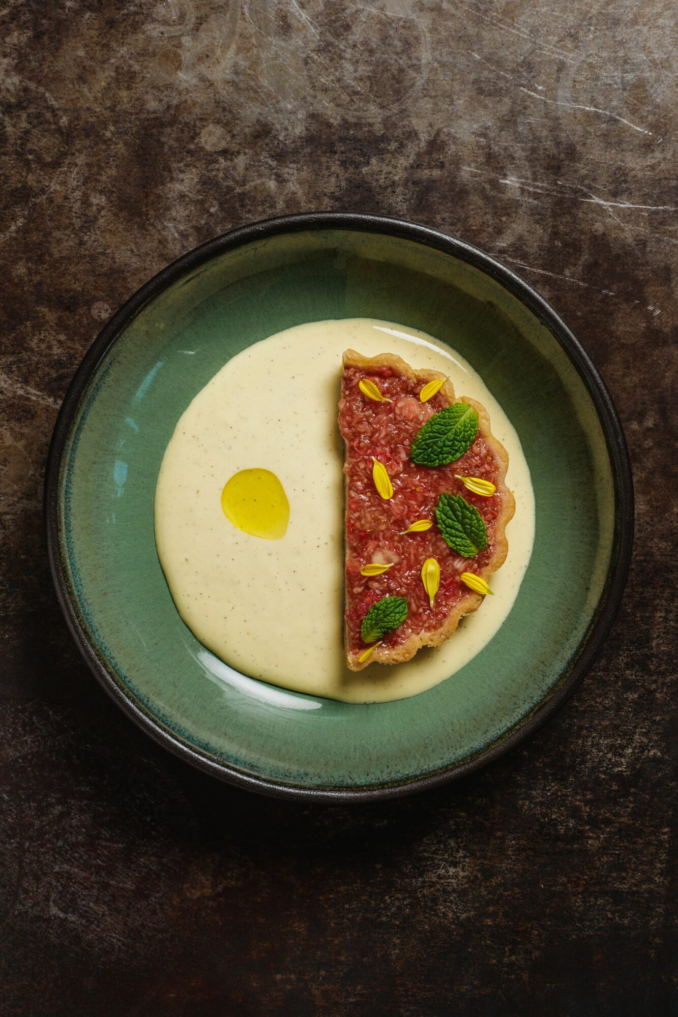 Creamy white soup in a bowl with a red topping garnished with green herbs, drizzled with oil, on a dark surface.