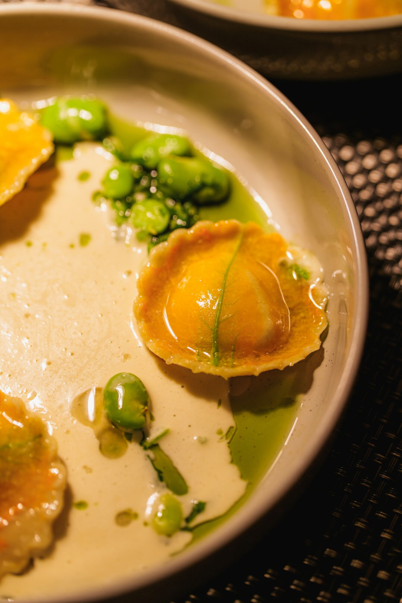 Close-up of a bowl with green peas, a halved yellow fruit, and a creamy liquid, on a black textured surface.