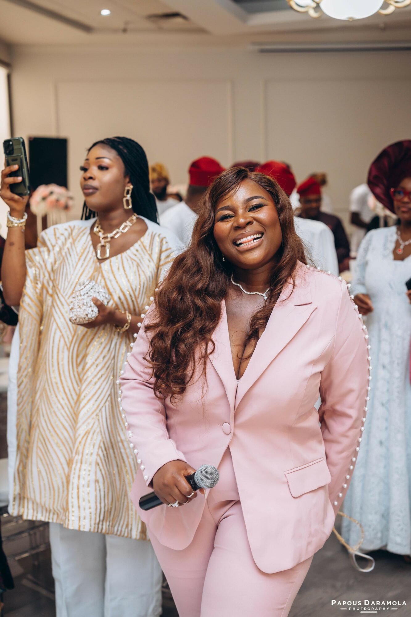 Smiling woman in pink suit holding a microphone, surrounded by women in traditional attire at an indoor event.