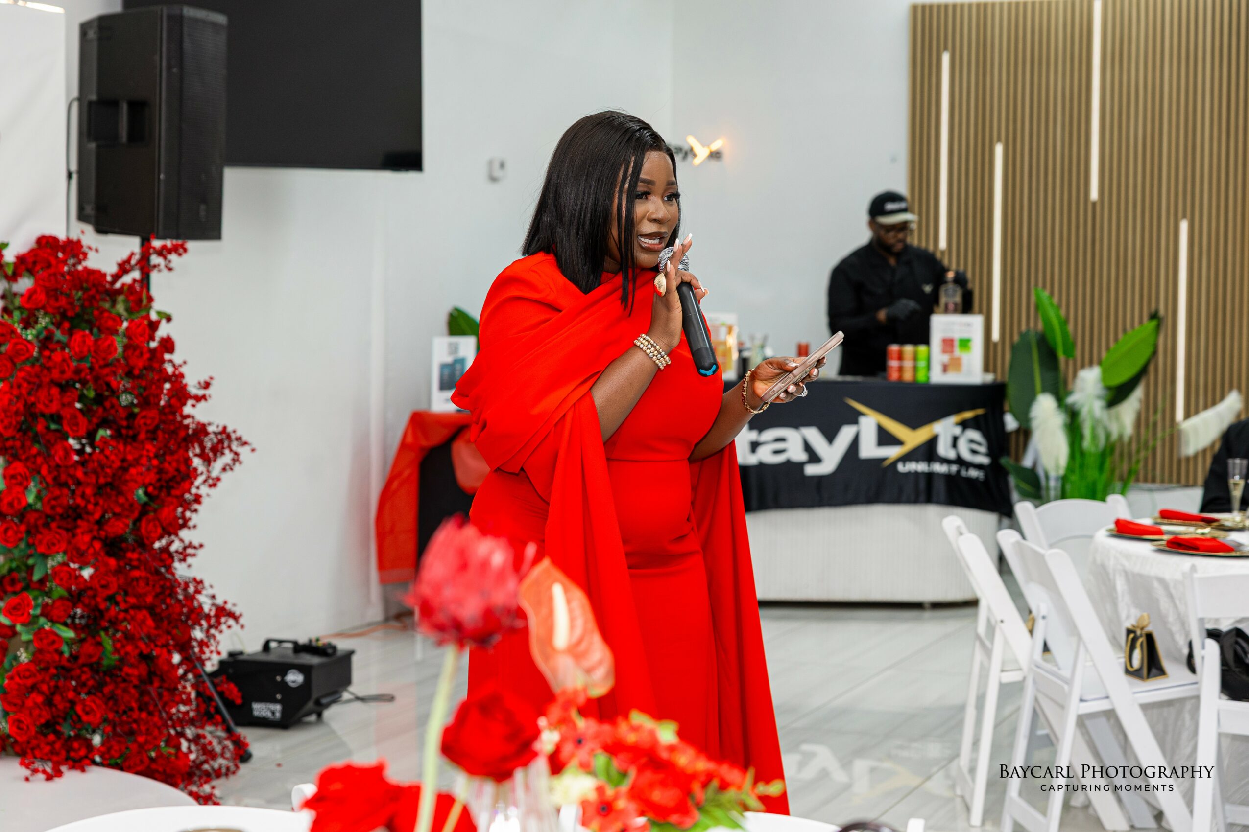 Woman in red dress speaking into microphone at event, with DJ and decorated tables in background.