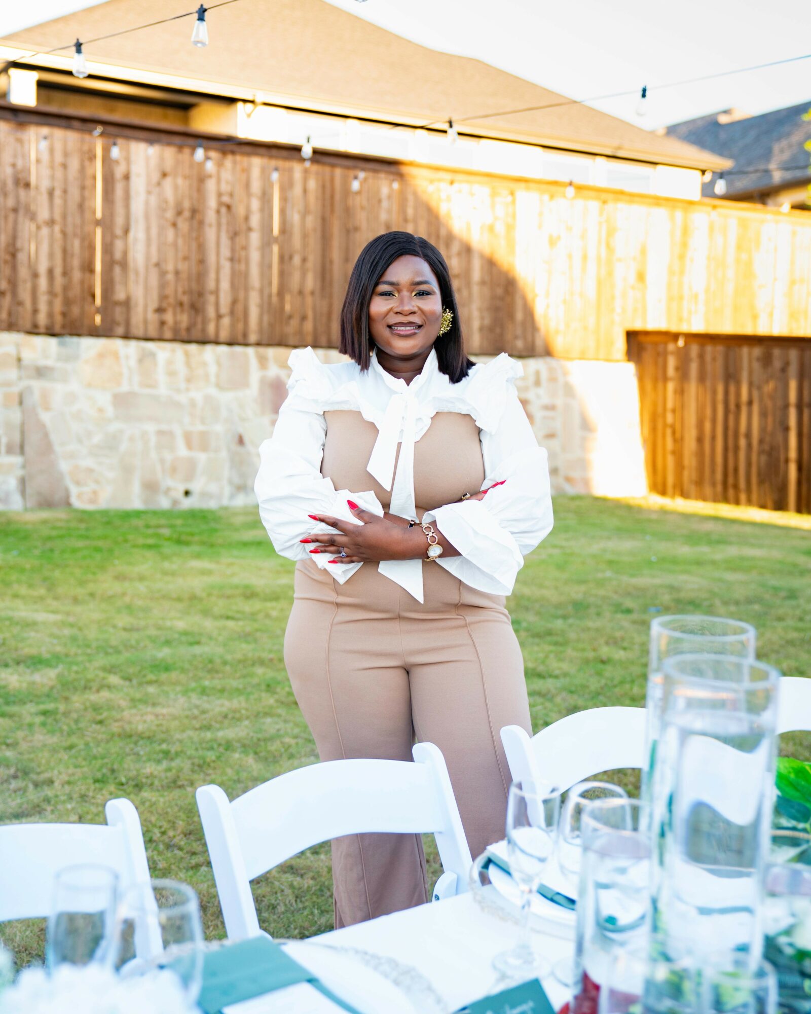 Woman standing outdoors in a backyard, smiling, with a table set with glasses and a pitcher in the foreground.