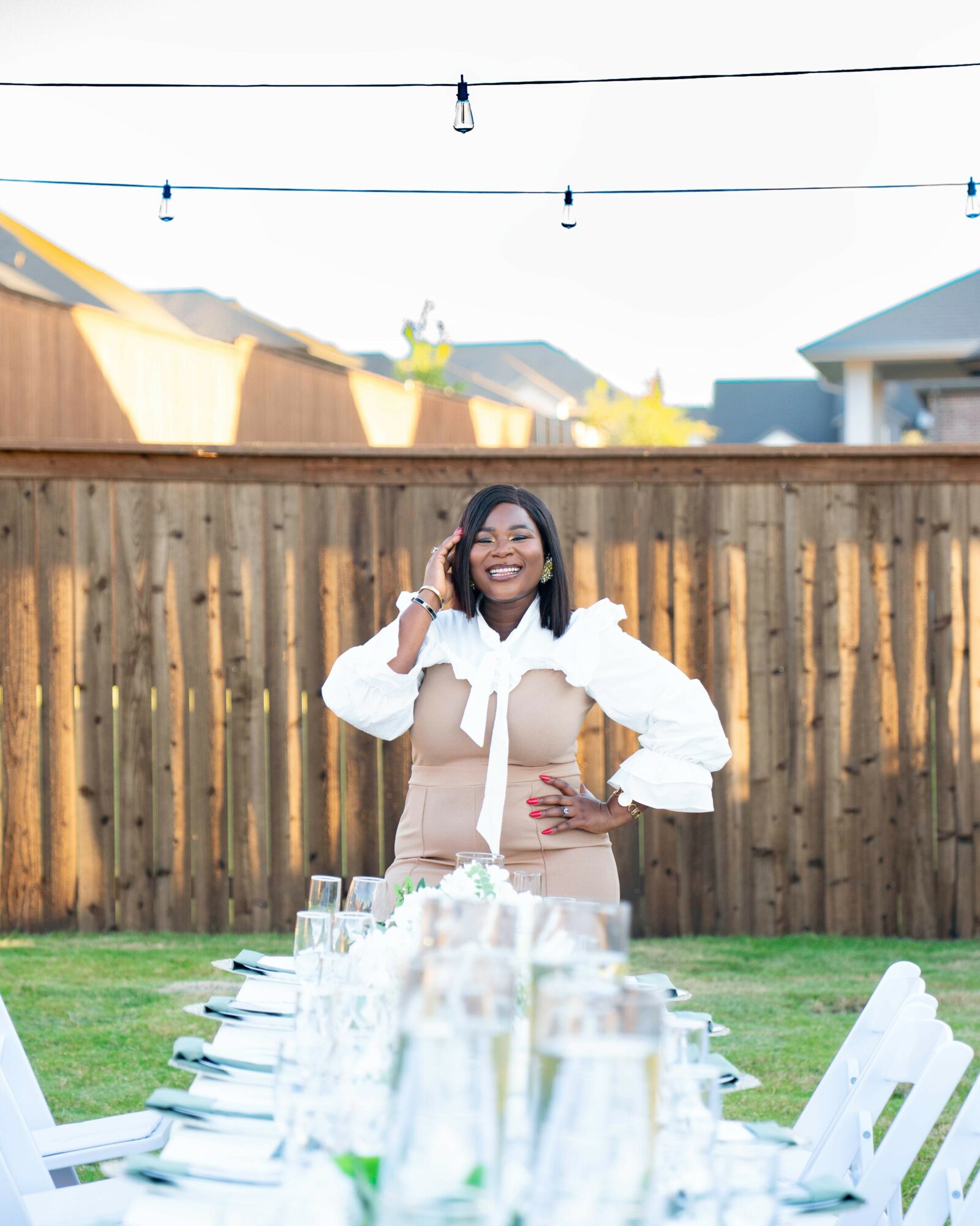 Woman smiling, talking on phone, standing outdoors behind a table with glasses, in front of a wooden fence.