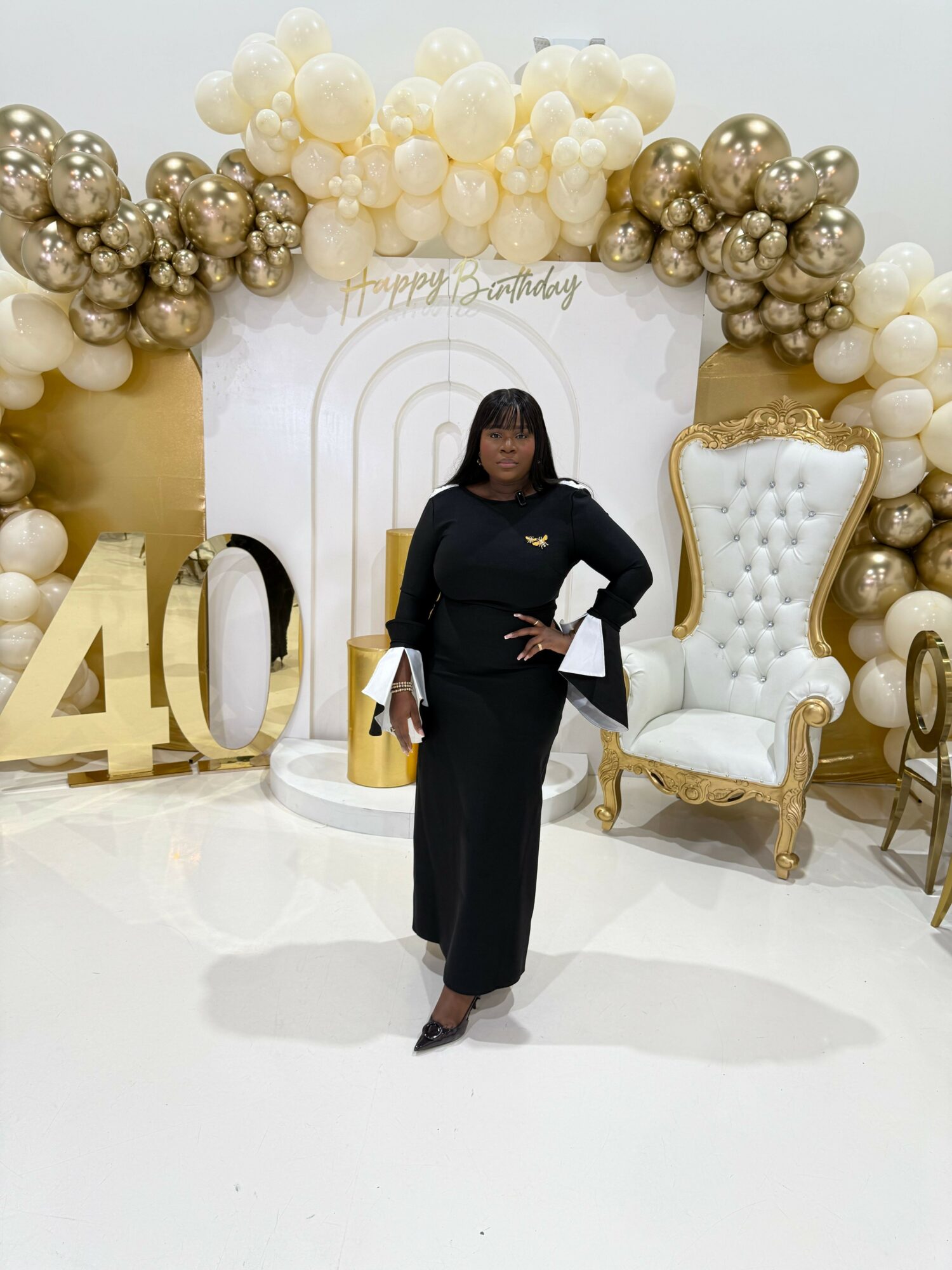 Woman in black dress standing in front of balloon arch and decorated backdrop with a white chair and large gold number 40.