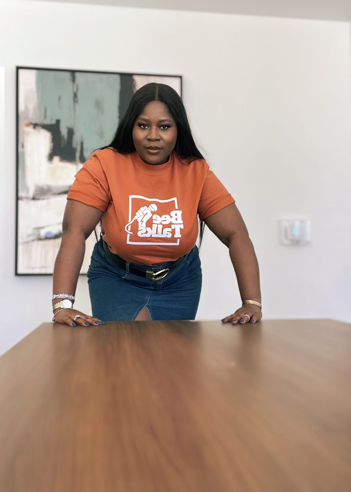 Woman with long dark hair wearing an orange T-shirt and blue jeans, leaning on a wooden table indoors.
