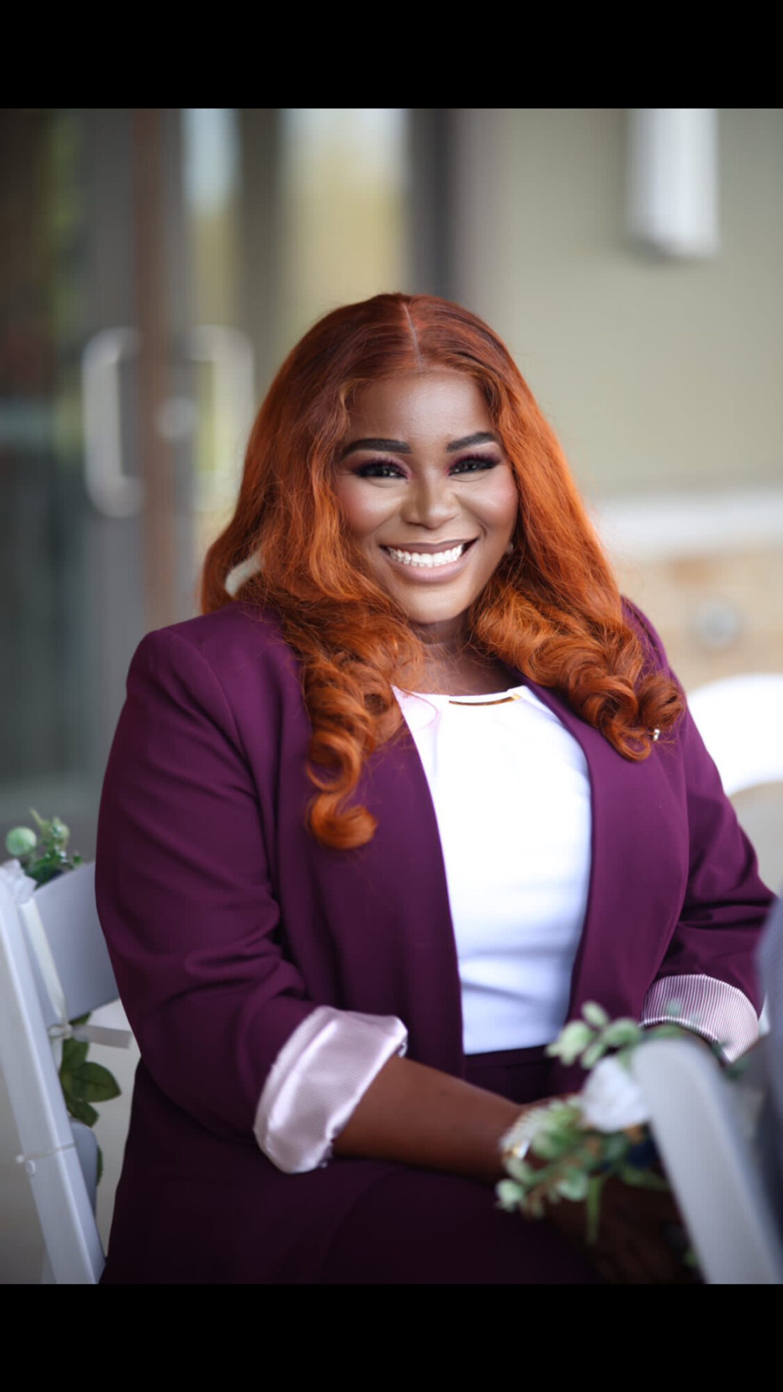 Smiling woman with orange hair in purple blazer and white top, sitting outdoors with greenery and white flowers.