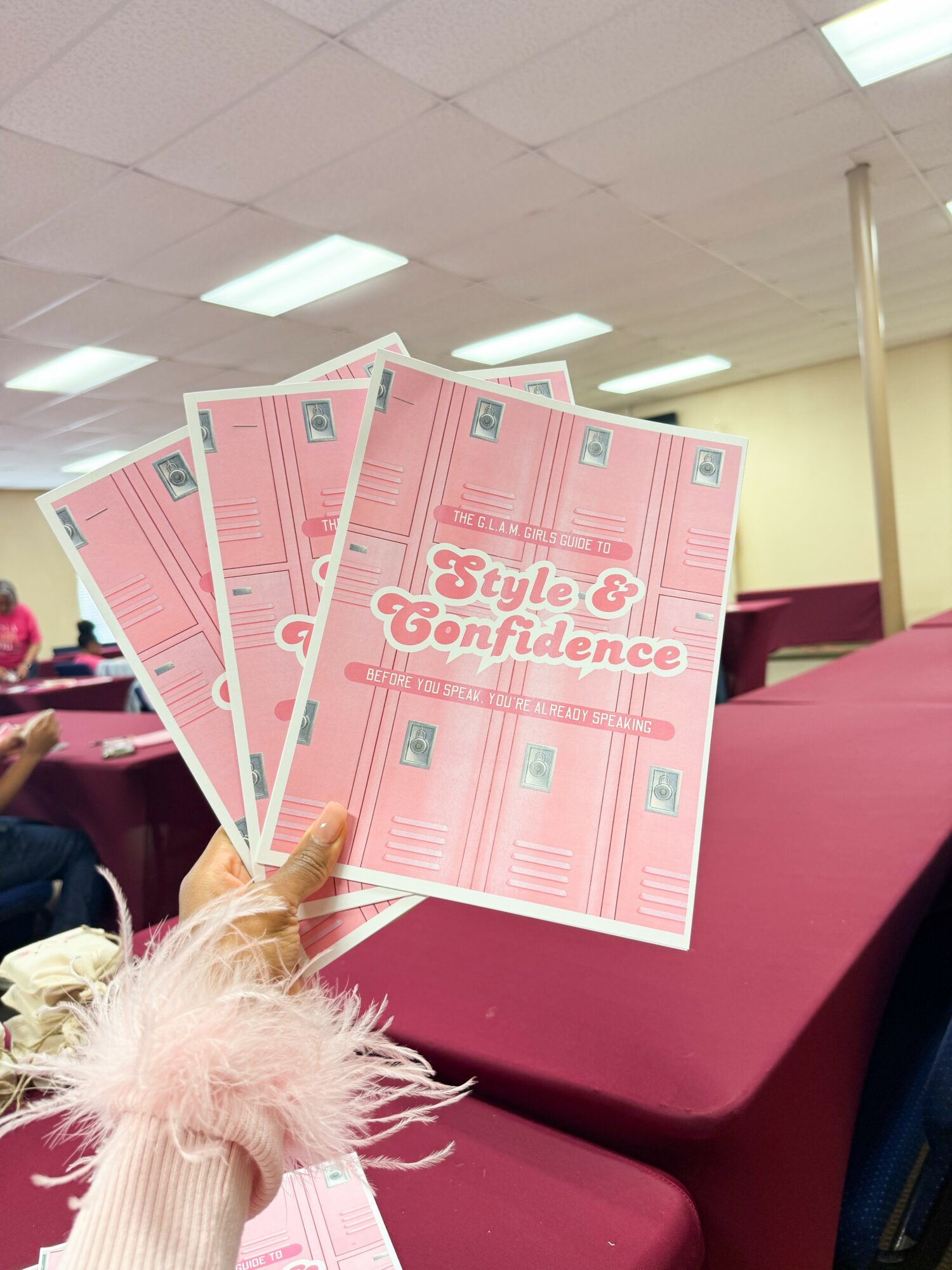 Person holding pink flyers with text 'Style & Confidence' in a room with tables and ceiling lights.