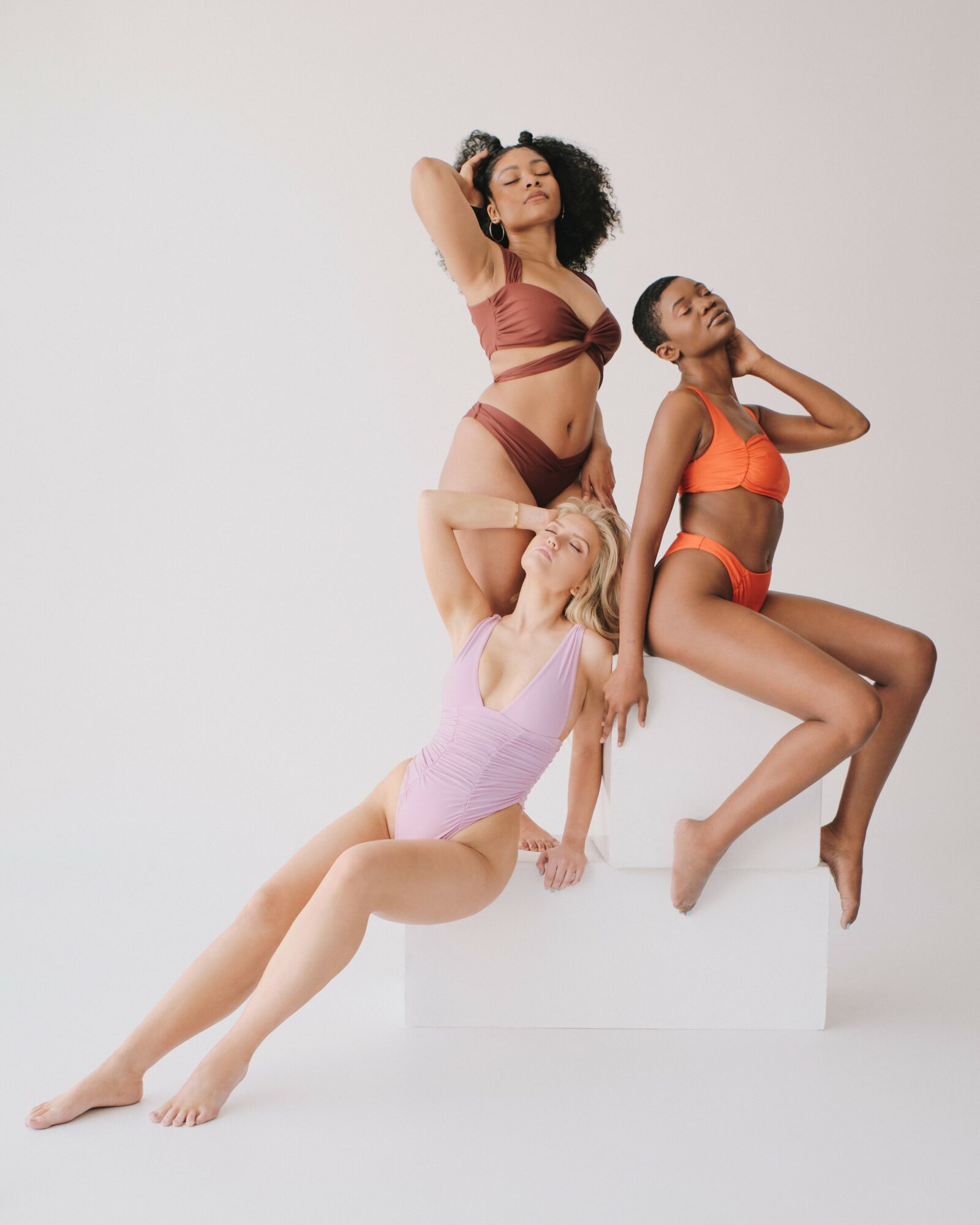 Three women in colorful lingerie pose on and around a white platform against a plain background.