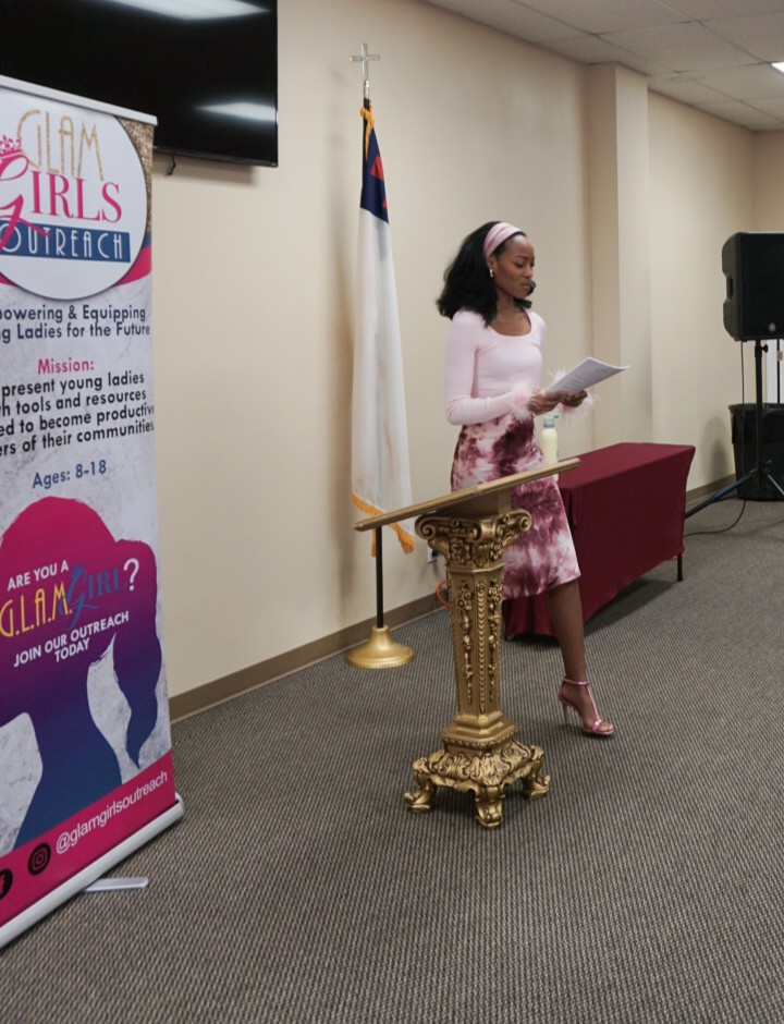 Woman standing at a decorated podium, reading from a paper, with a flag and a banner nearby.