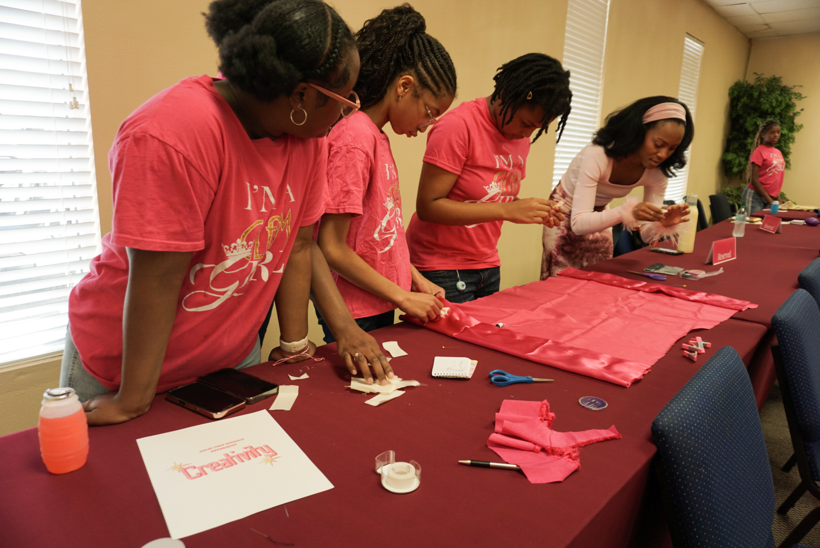 Five women in pink shirts work together on a craft project at a table with pink fabric and supplies.