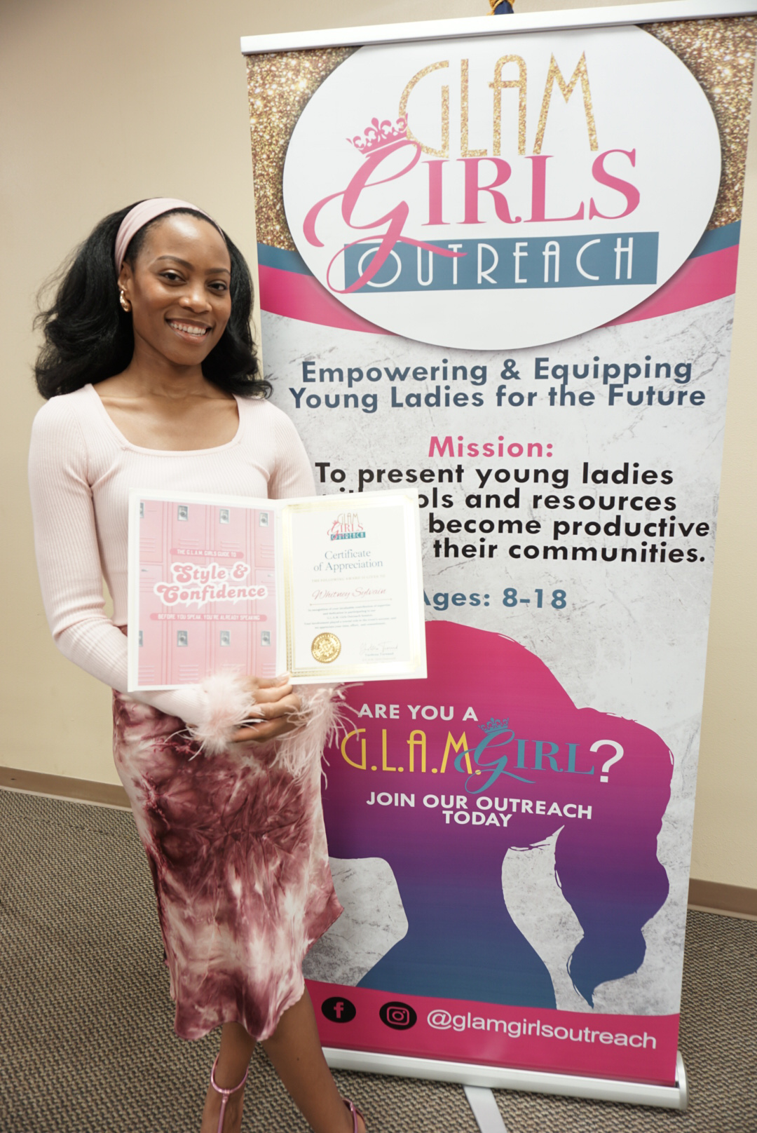 Young girl in pink and white outfit standing next to a banner for G.L.A.M Girls Outreach, smiling.