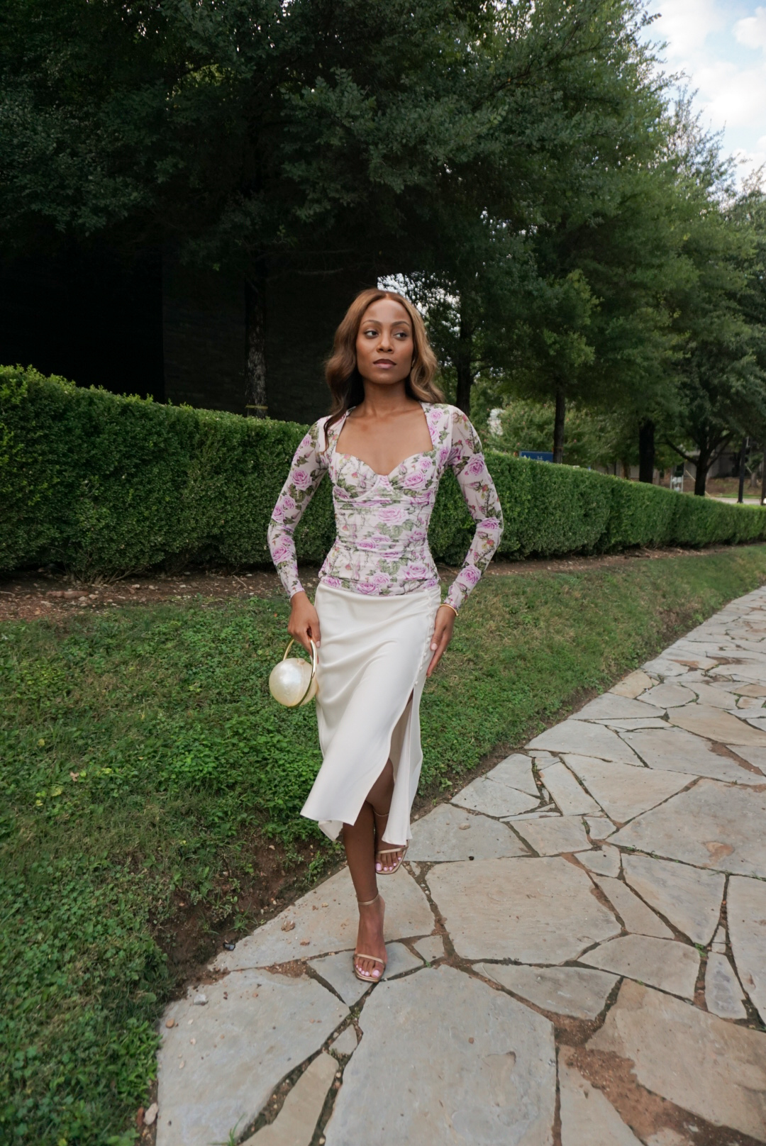 Woman in floral top and white skirt walking on stone path with greenery background.