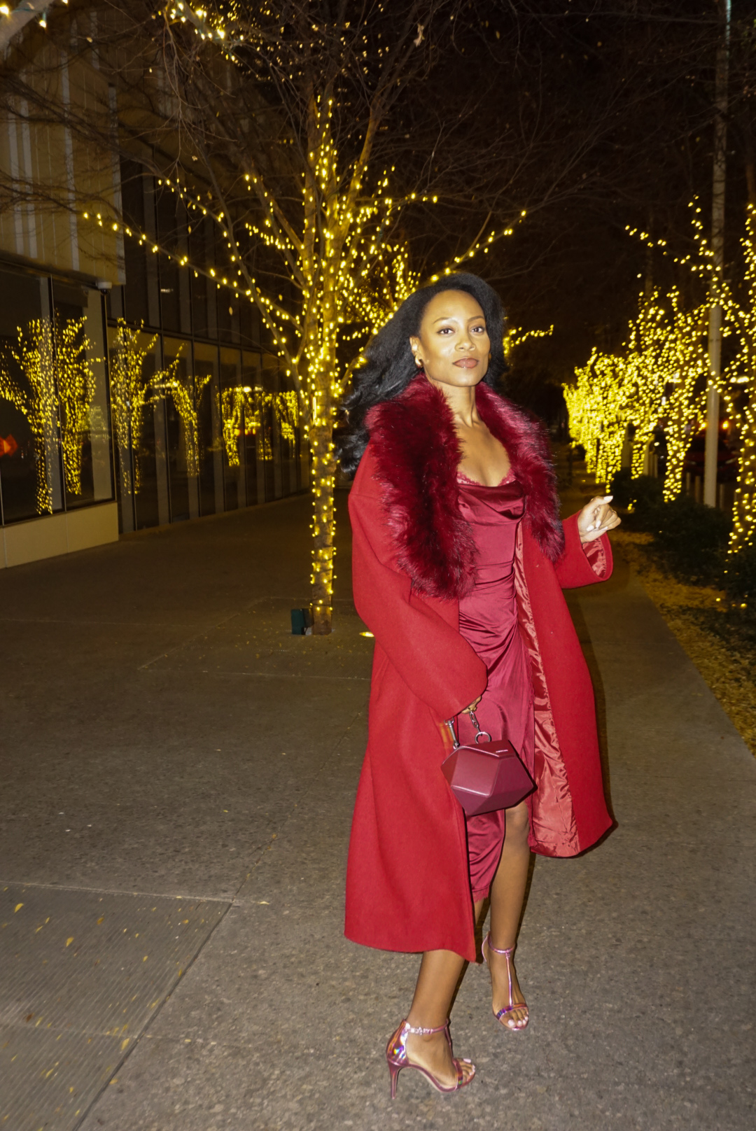 Woman in red coat and dress standing outdoors at night with illuminated trees in background.